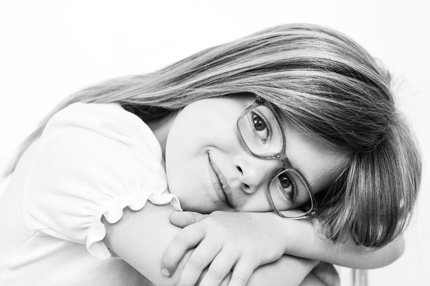 Black and white portrait of a smiling young girl wearing glasses, resting her arms and chin on a surface.