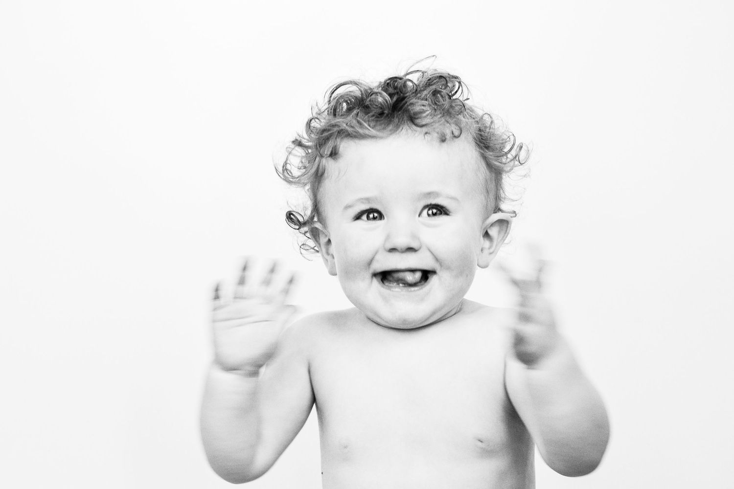 Black and white portrait of a happy baby with curly hair raising both hands.