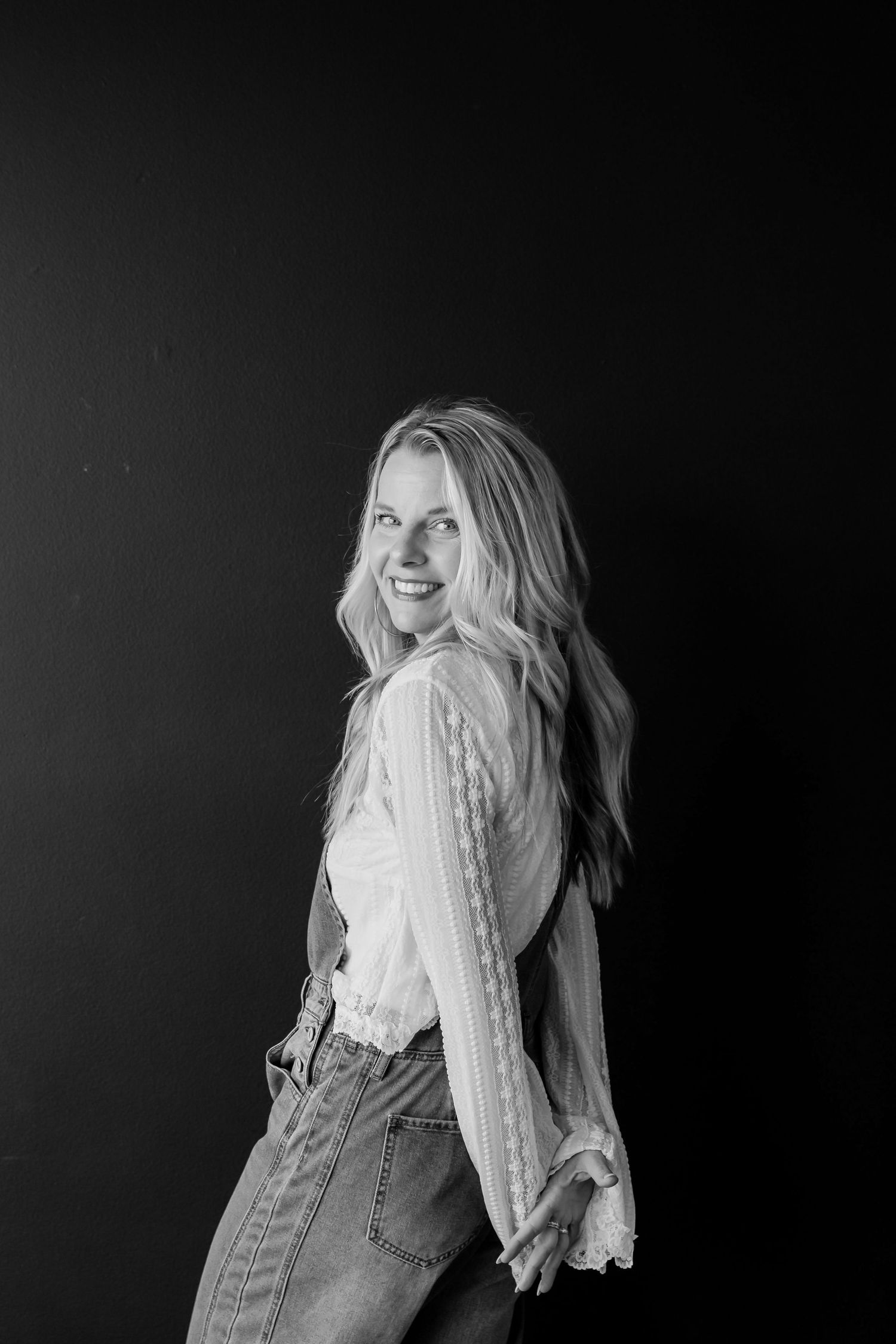 Blonde woman in lace blouse and jeans smiles over shoulder in black and white portrait.