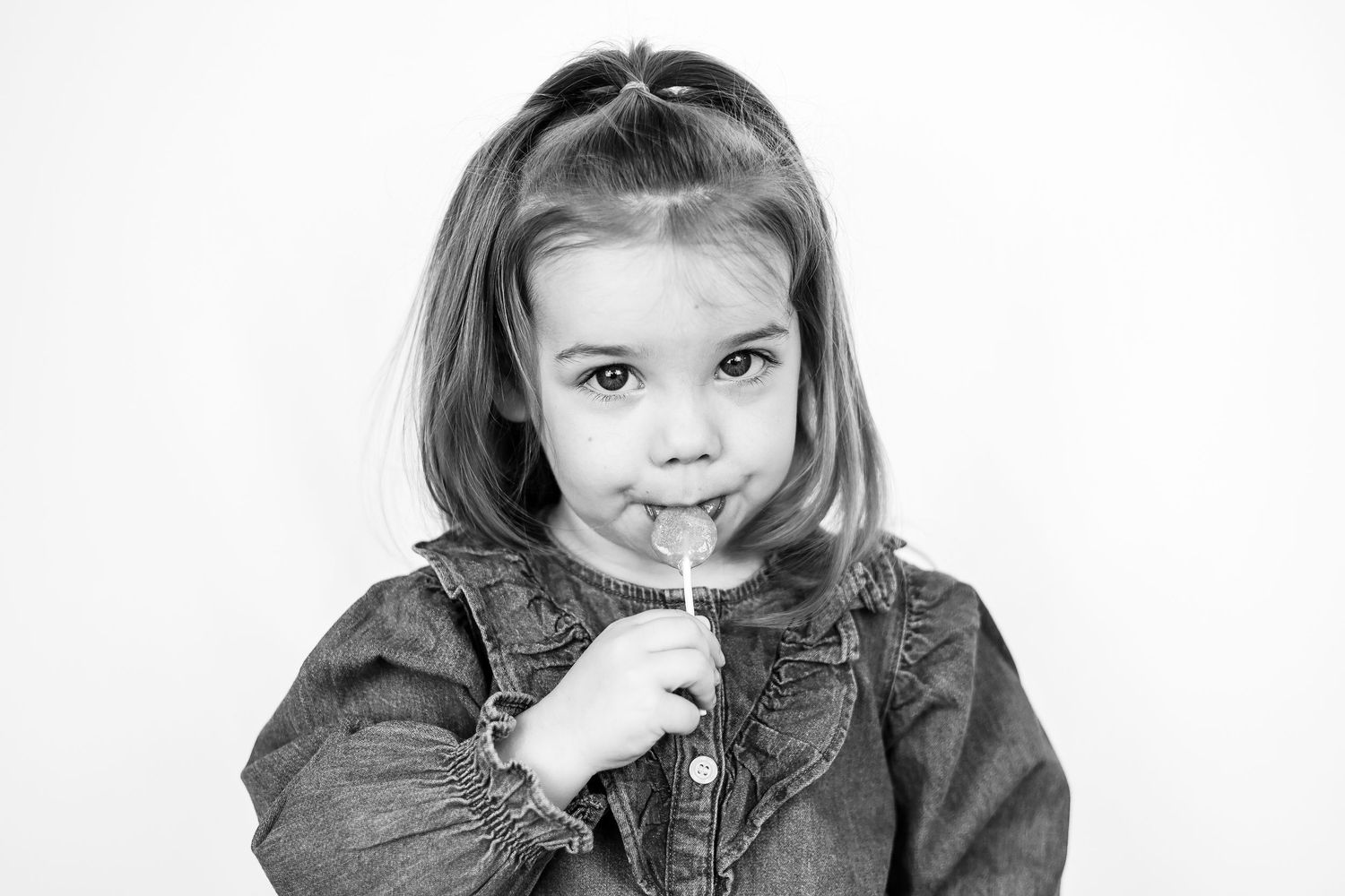 Cute little girl in denim jacket holds a lollipop in a black and white portrait.