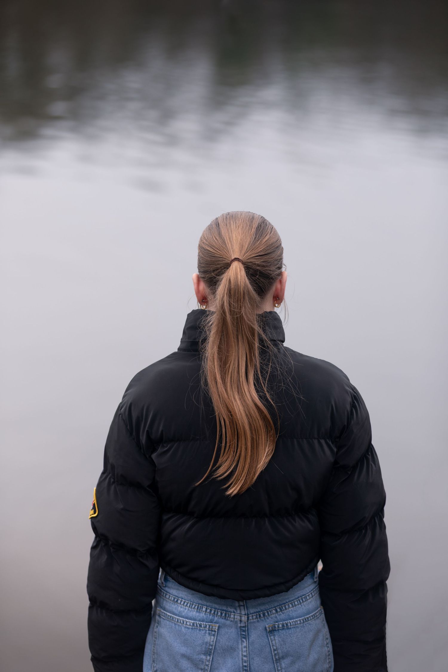 Young girl with ponytail in black puffer jacket stands facing calm misty lake.

Keyingham