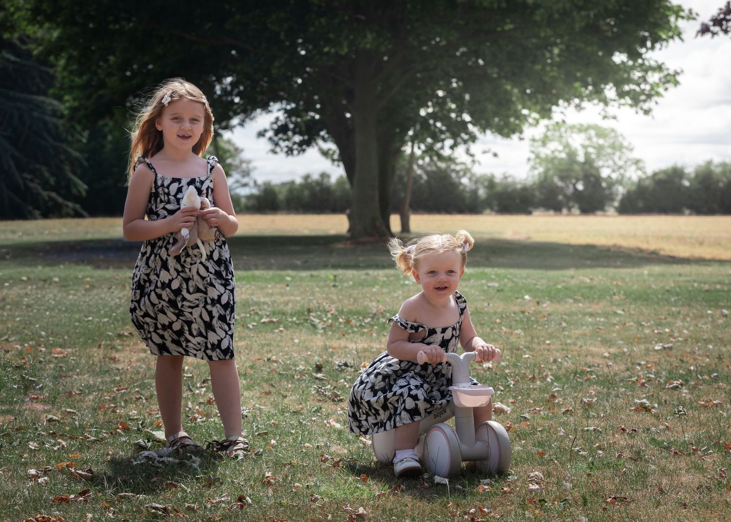 Two young girls in matching black and white dresses playing outdoors in a sunny park with large trees.