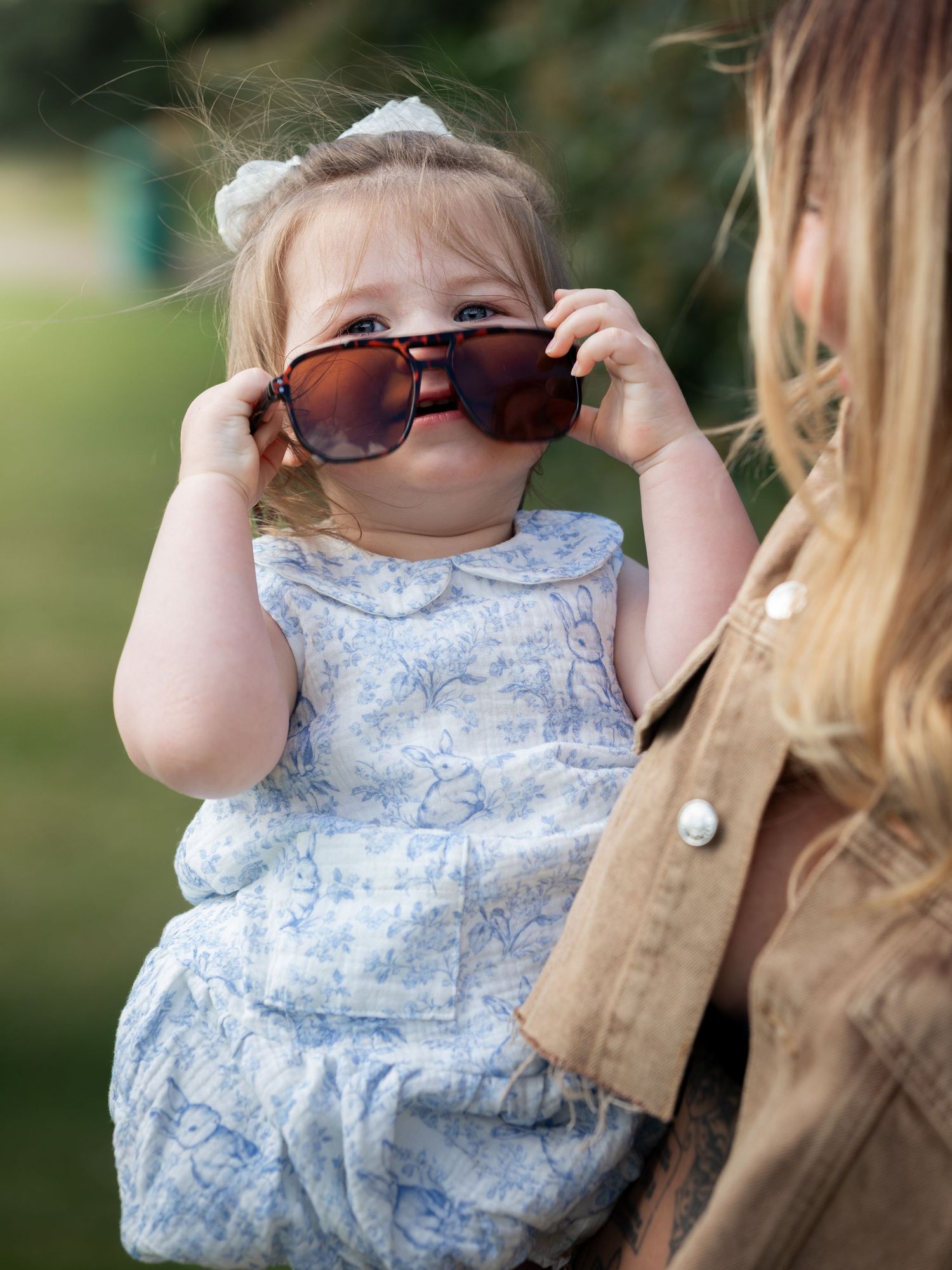 Adorable toddler girl in floral dress trying on oversized sunglasses while being held by her mother outdoors.
East Park Hull