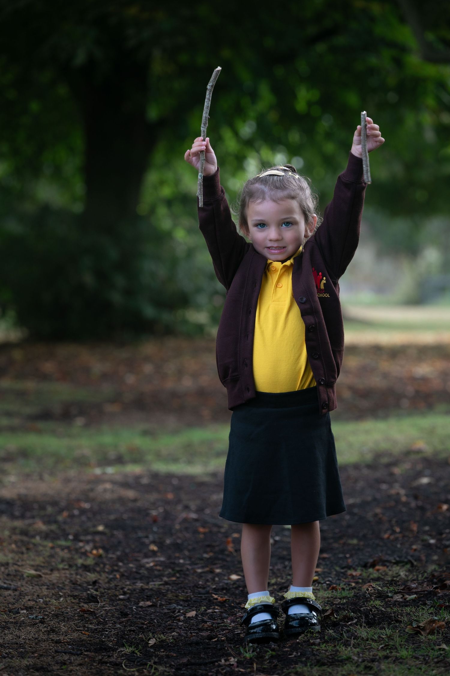 Young girl in school uniform raises sticks above her head while standing in a park.