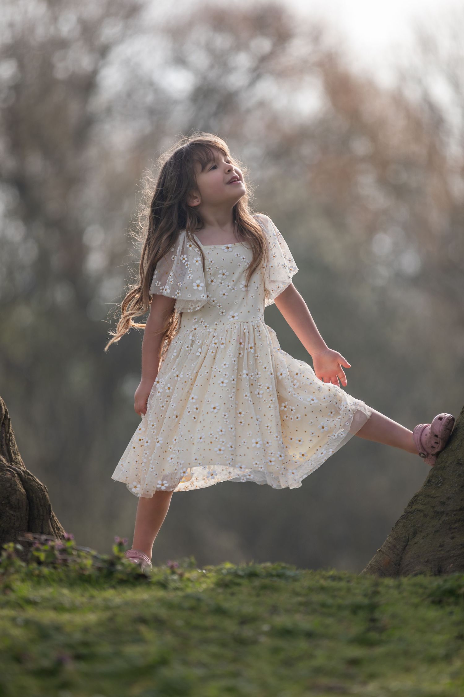 Young girl in white floral dress poses outdoors among trees on green grass in soft natural light.