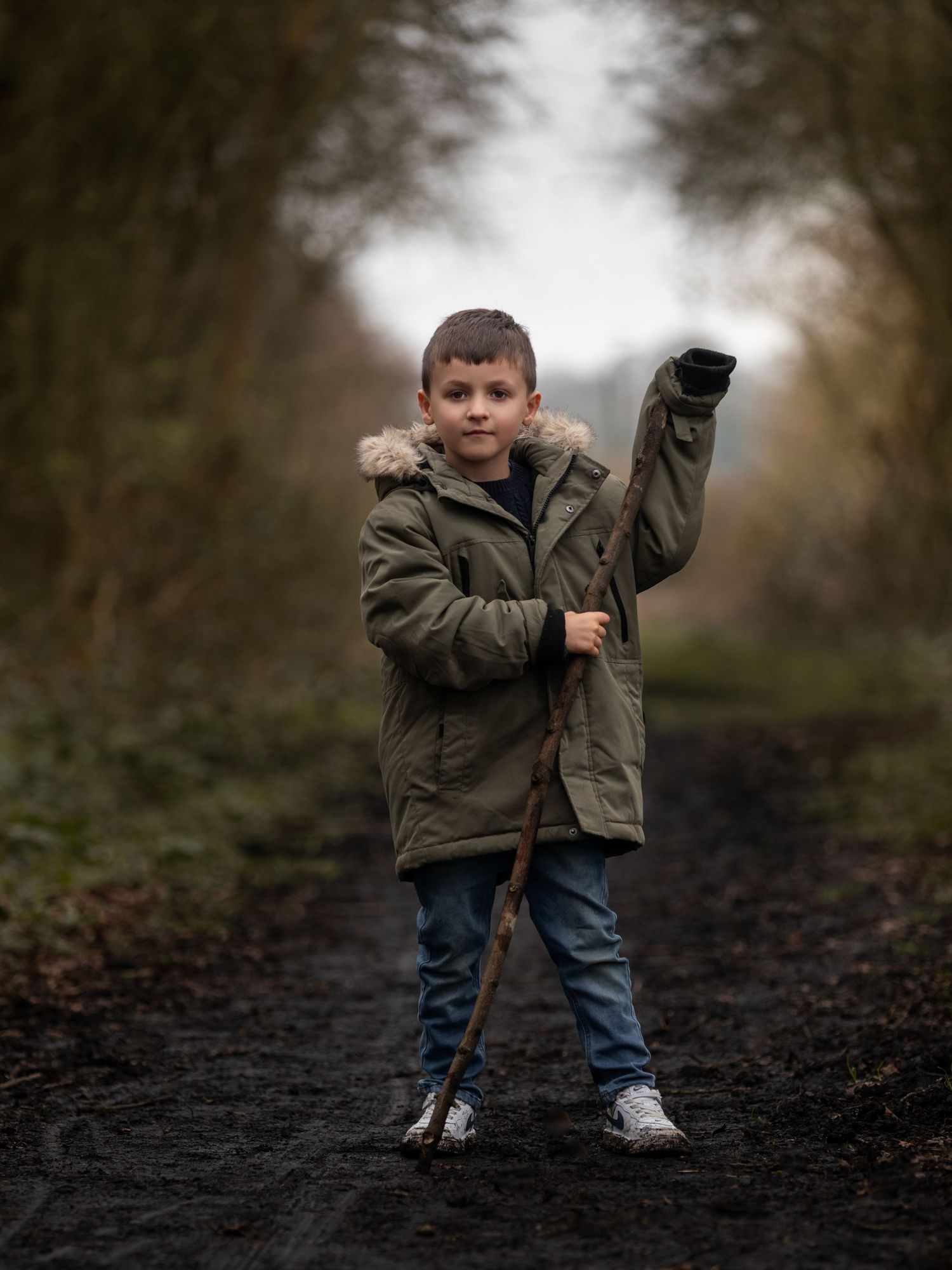Young boy in olive parka holding a stick on a muddy forest path, waving at the camera.