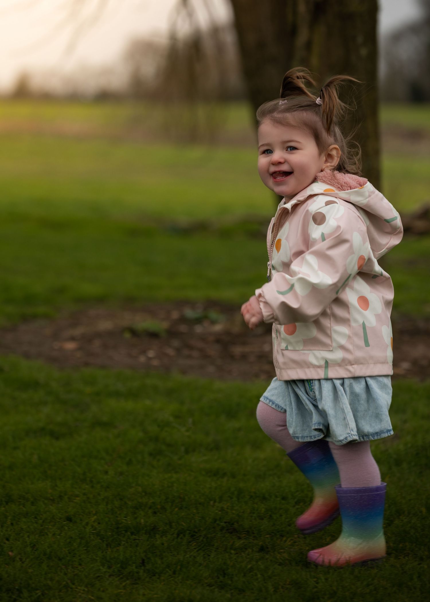 Happy toddler girl in polka dot jacket and rainbow boots running on green grass.