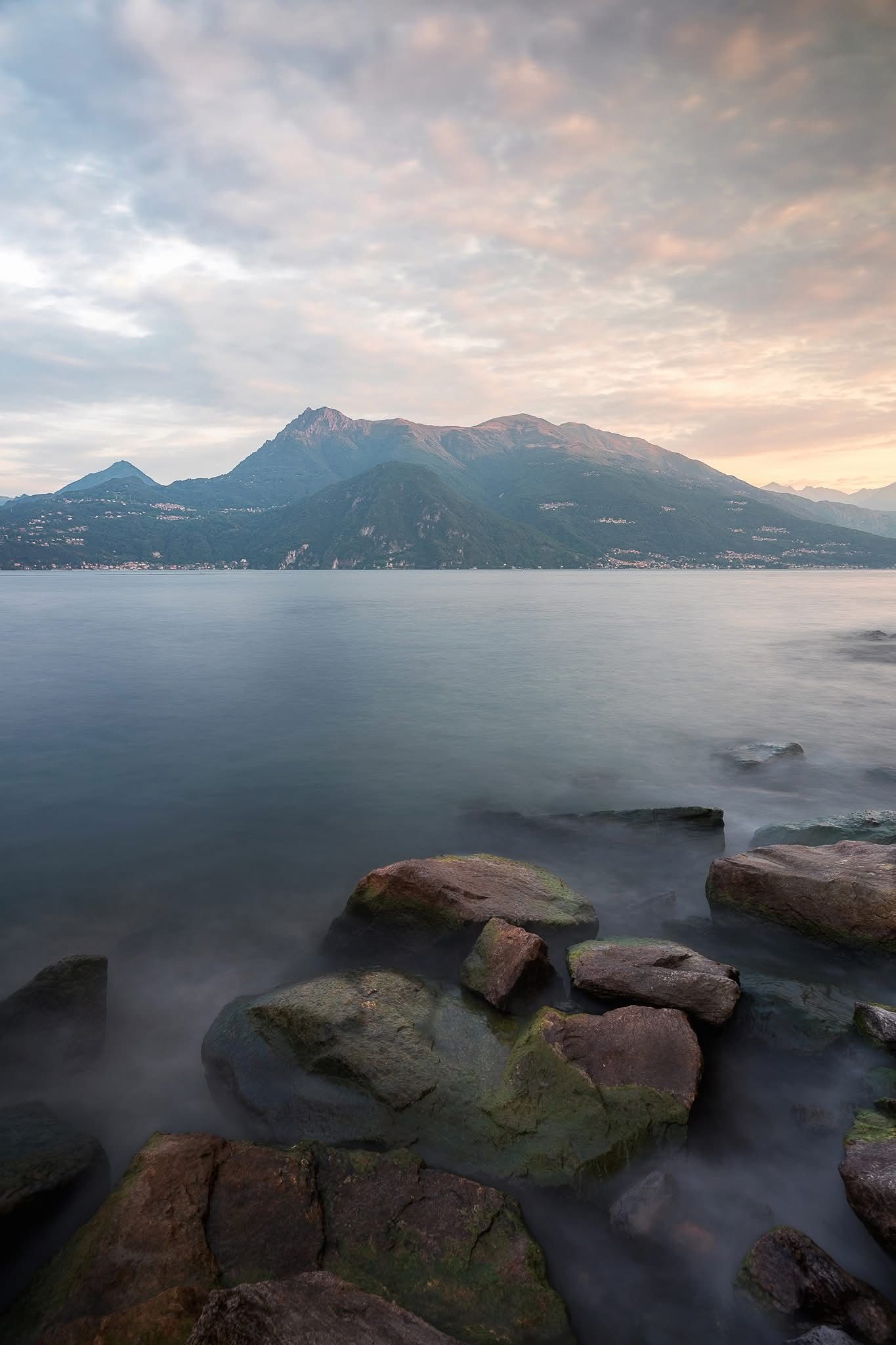 Misty mountain lake at sunset with mossy rocks in the foreground and pink clouds.

Varenna Italy