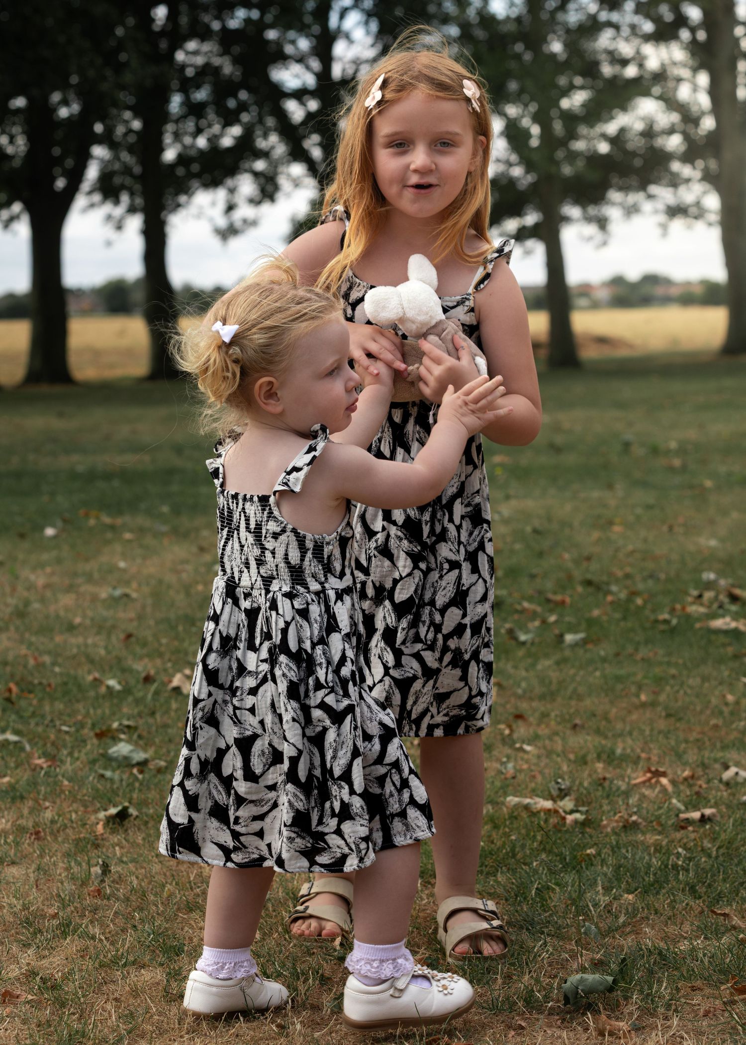 Two young blonde sisters in matching black and white dresses share a stuffed animal toy outdoors in a park.
