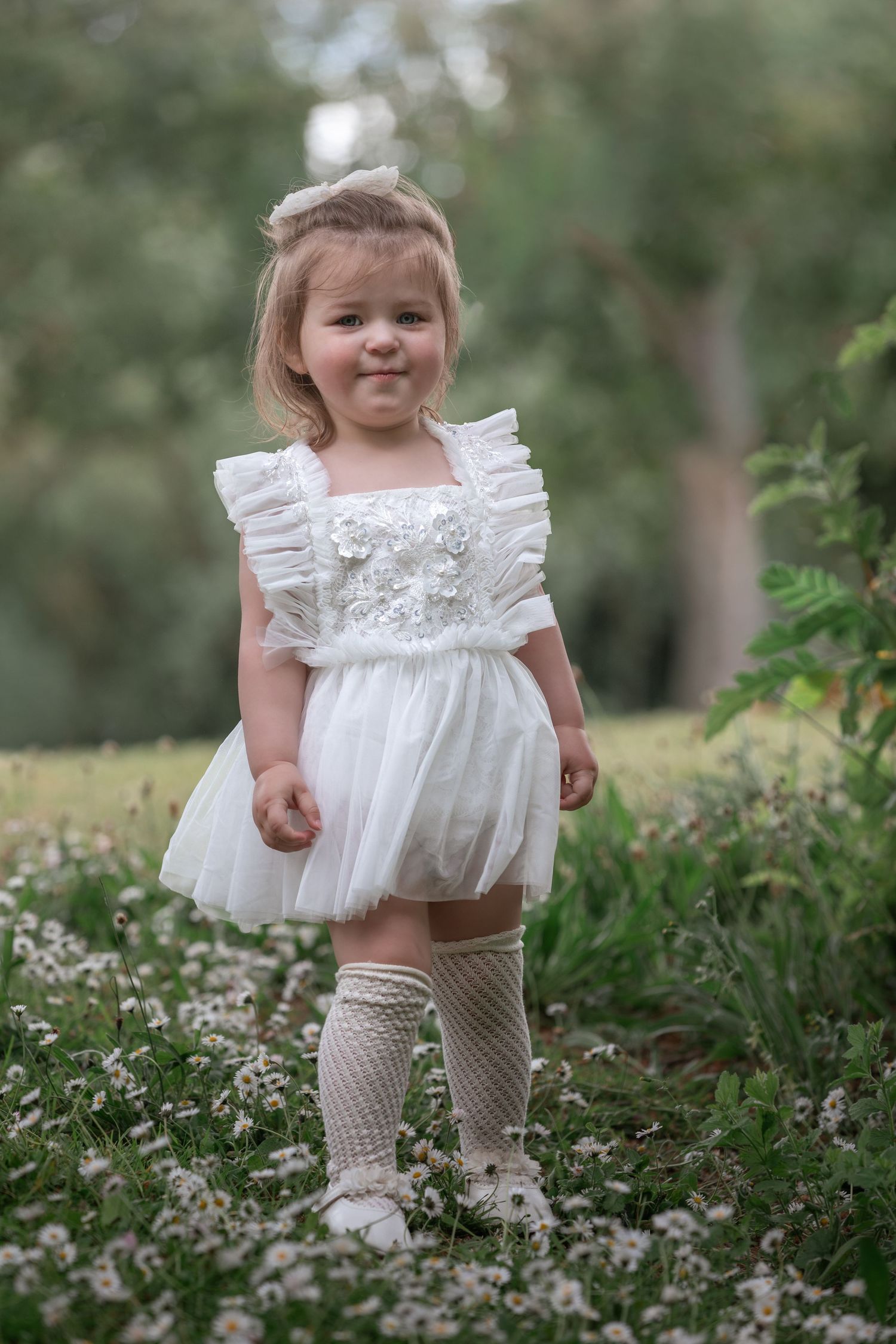 Toddler girl in white ruffle tulle dress and knee socks standing among wildflowers in a lush green garden.

East Park Hull