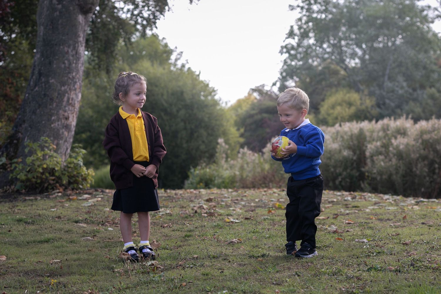 Two young children playing outdoors on grass, girl in school uniform and boy holding colorful ball near trees.