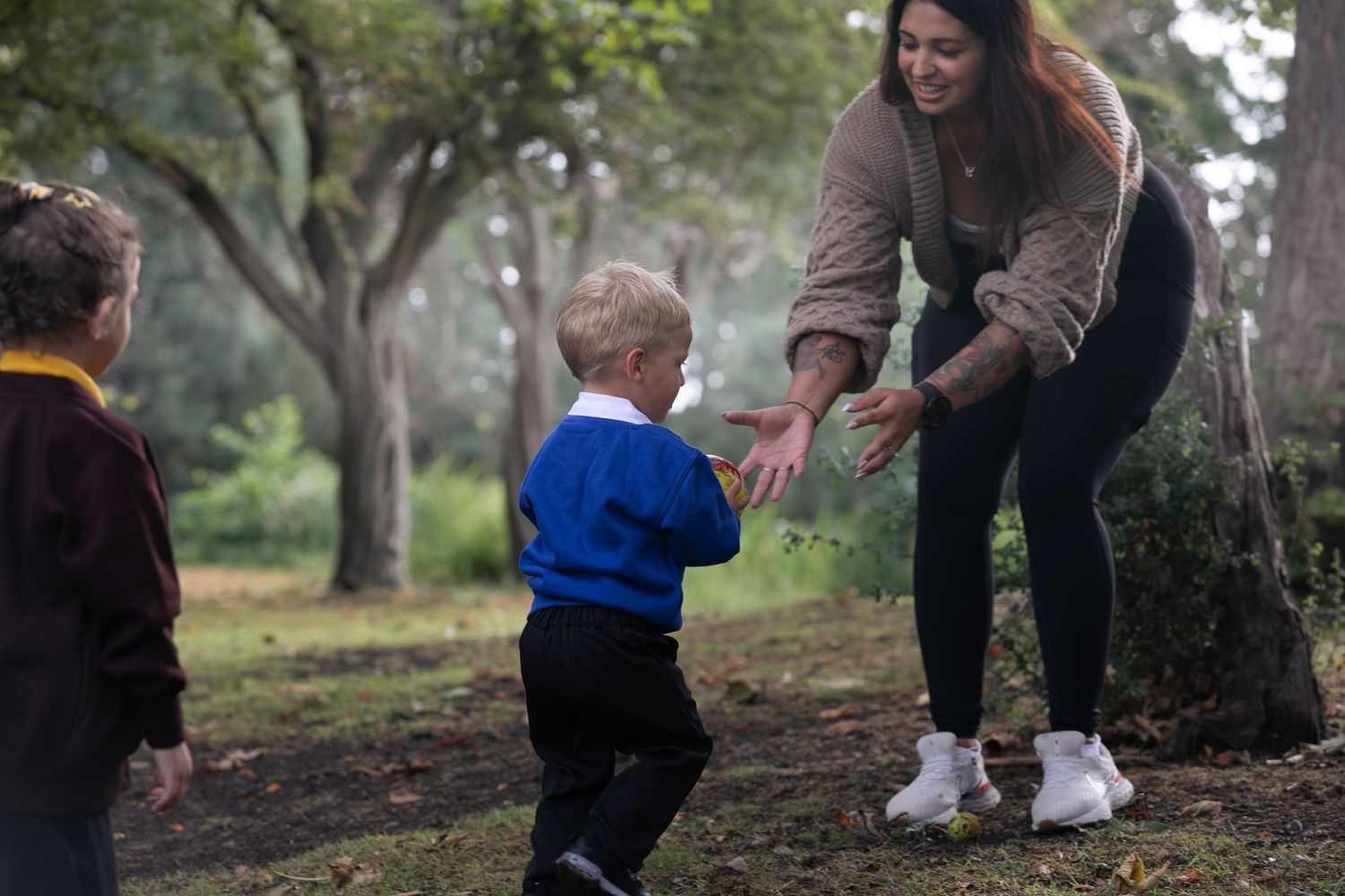 Woman leaning forward passing a ball to a young boy in blue school uniform outdoors in a misty park.