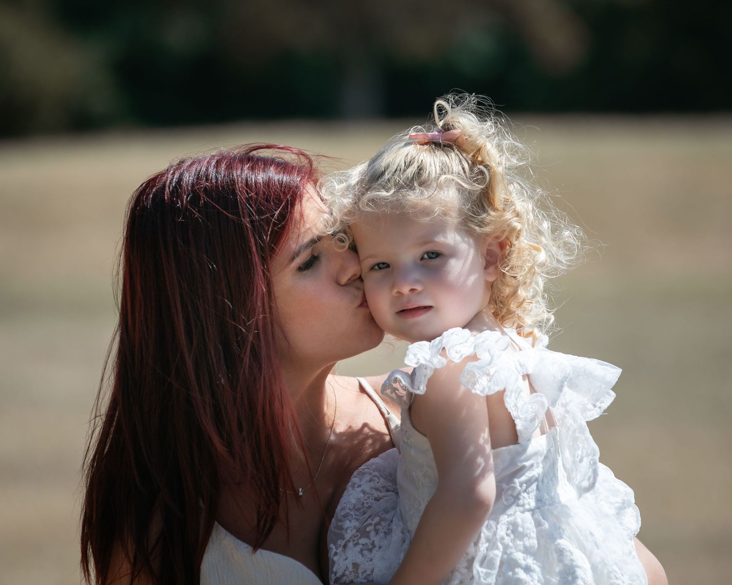 A mother with red hair kisses her curly-haired toddler girl in a white dress outdoors.