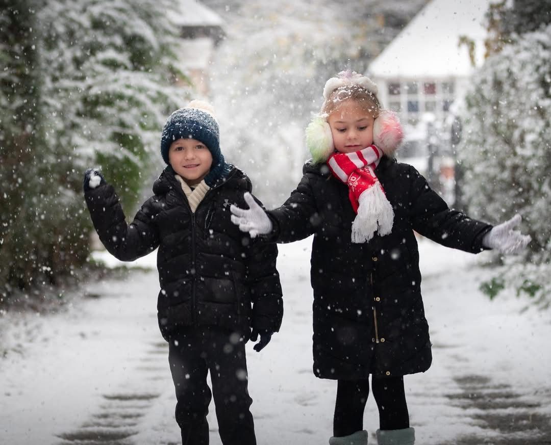 Two children playing in snowfall outdoors, wearing winter coats and scarves, joyfully catching snowflakes in a snowy garden.