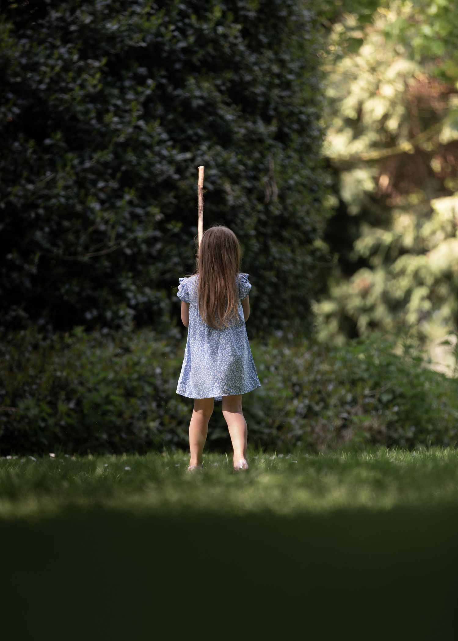 A young girl in a blue dress stands barefoot on grass, holding a wooden stick up toward lush green trees.