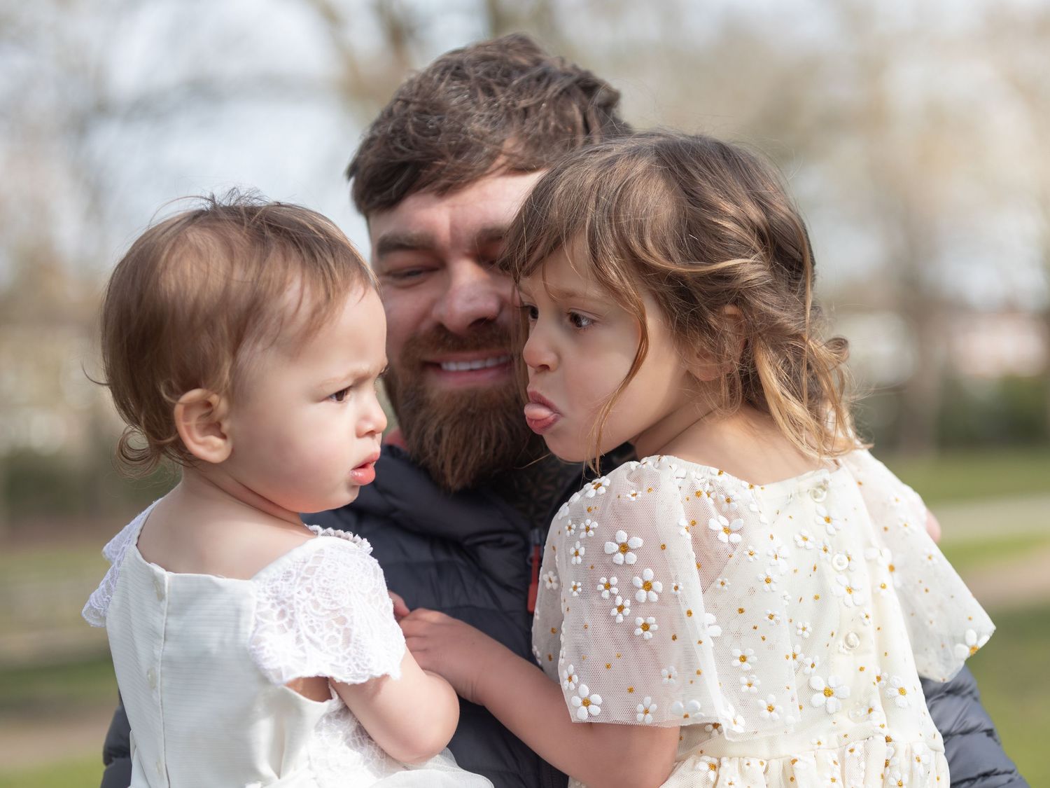 A bearded father tenderly holds two young daughters in white dresses outdoors in a sunny park setting.

East Park Hull