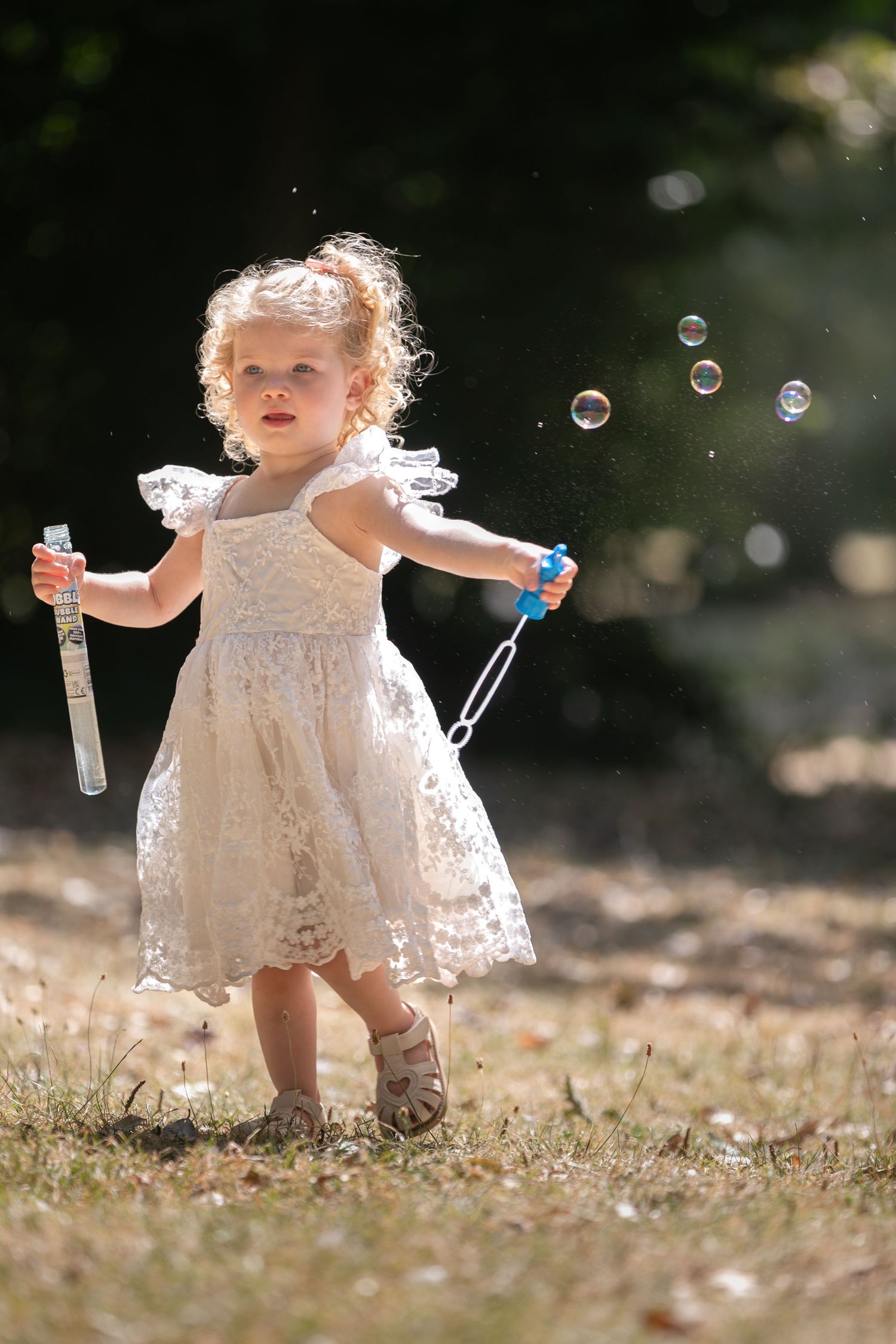 A curly-haired toddler girl in a white lace dress blows soap bubbles outdoors in a sunlit garden.