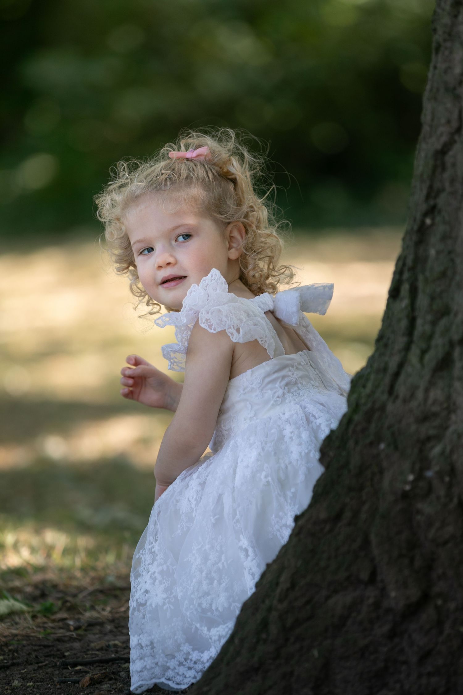 Young girl in white lace dress with pink bow leans against tree trunk in sunlit park setting.