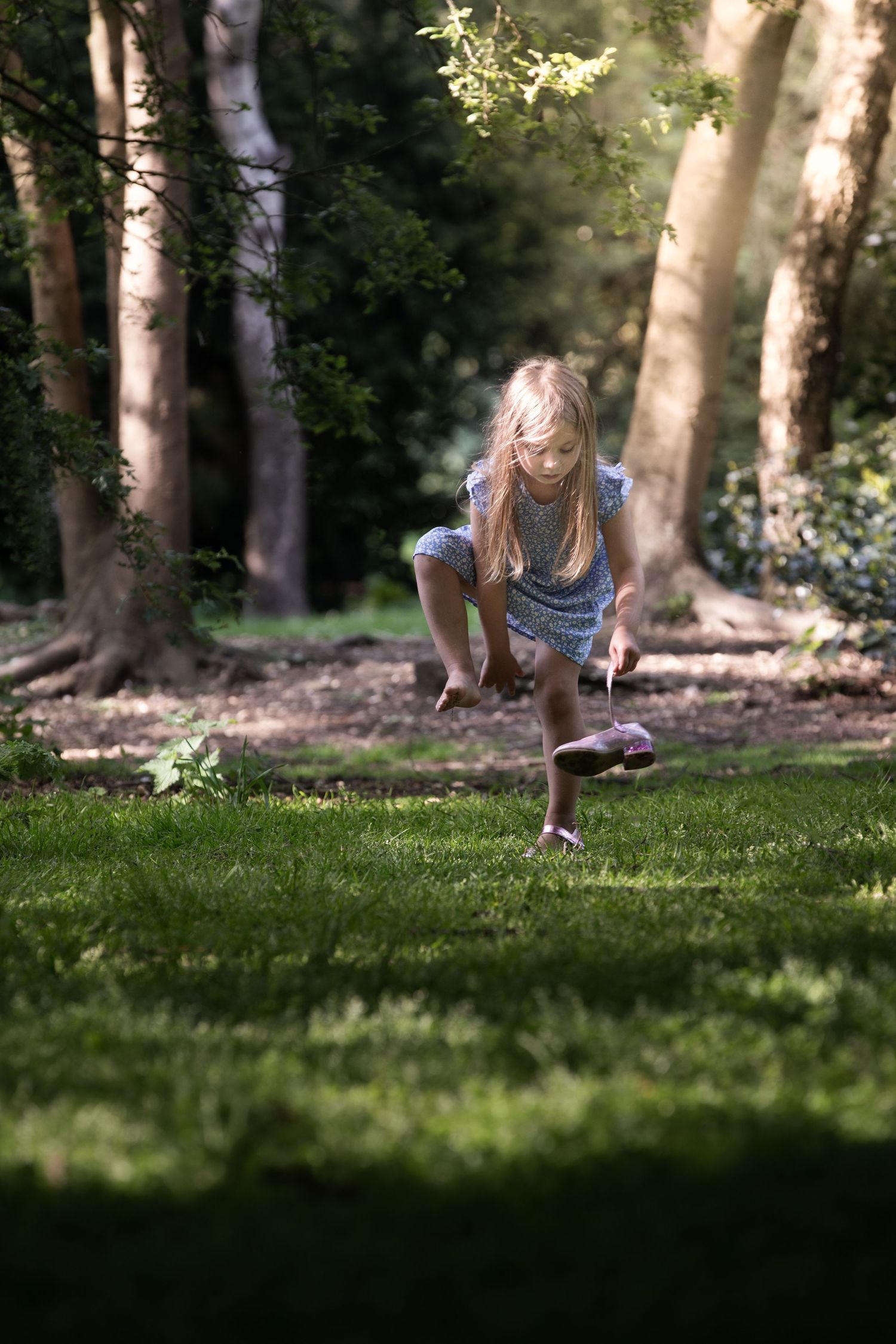 A young blonde girl in a blue dress bends down to pick something up while playing in a sunlit forest.