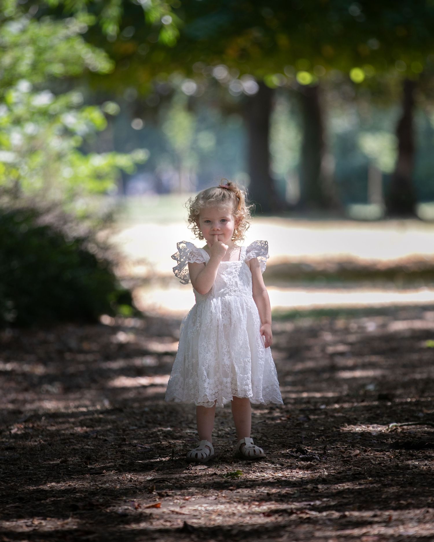 A toddler girl in a white lace dress stands on a tree-lined path, touching her face with one hand.