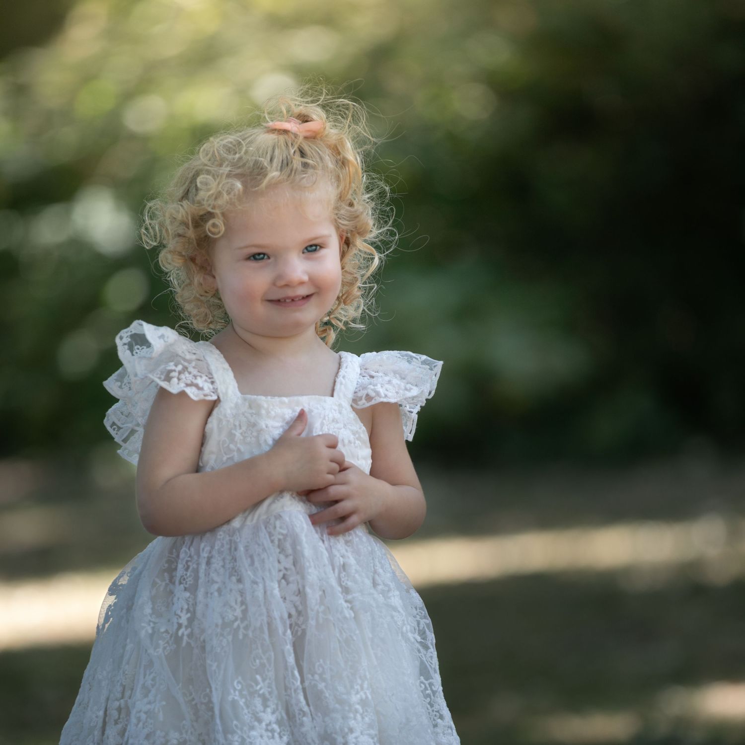 A smiling toddler girl with curly blonde hair wearing a white lace dress stands outdoors in a garden setting.