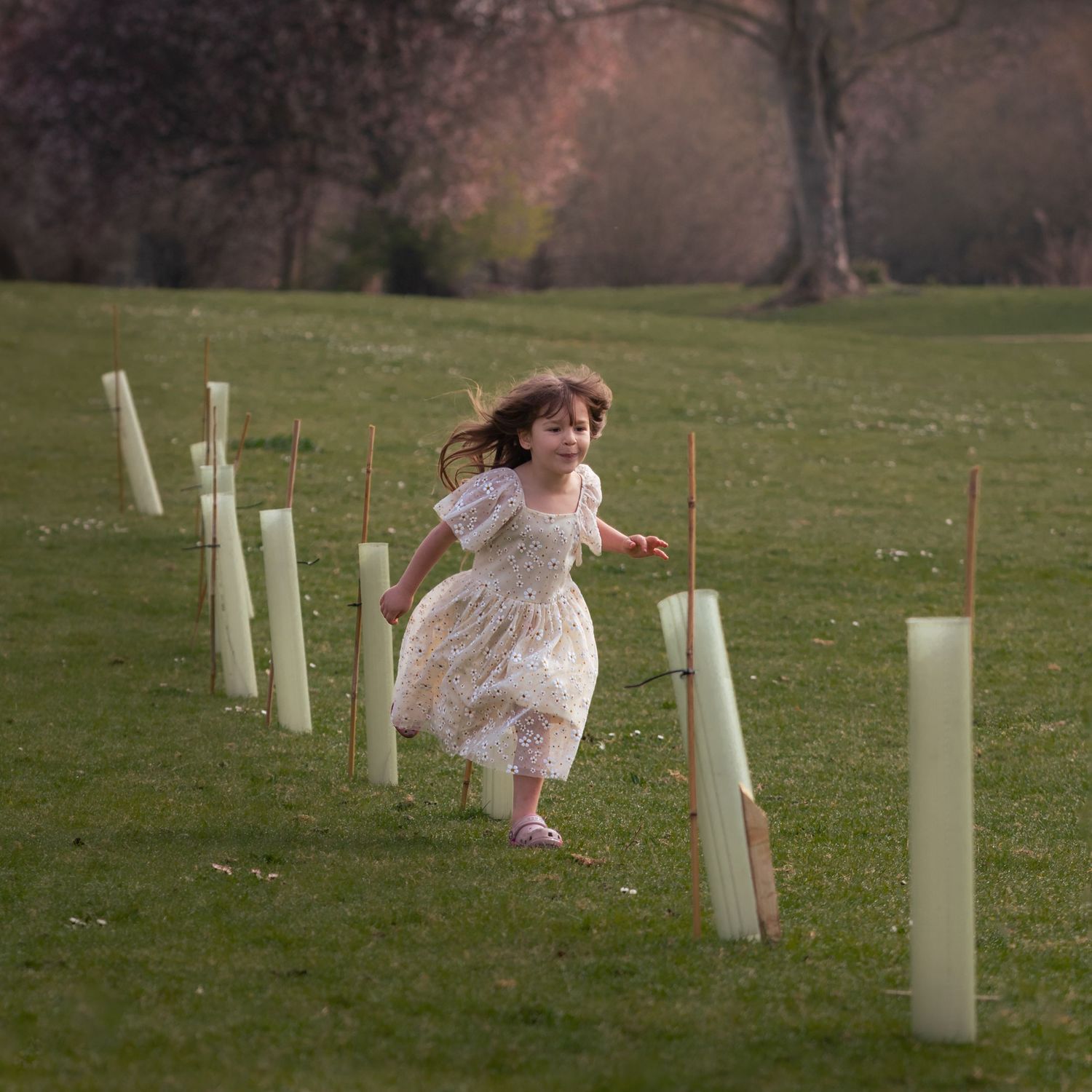 A young girl in a white lace dress runs joyfully through a green park lined with tree saplings in protective tubes.