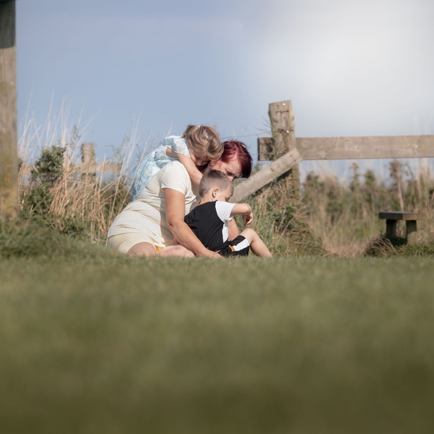A mother tenderly kisses her young child while sitting on grass near a wooden fence on a sunny day.