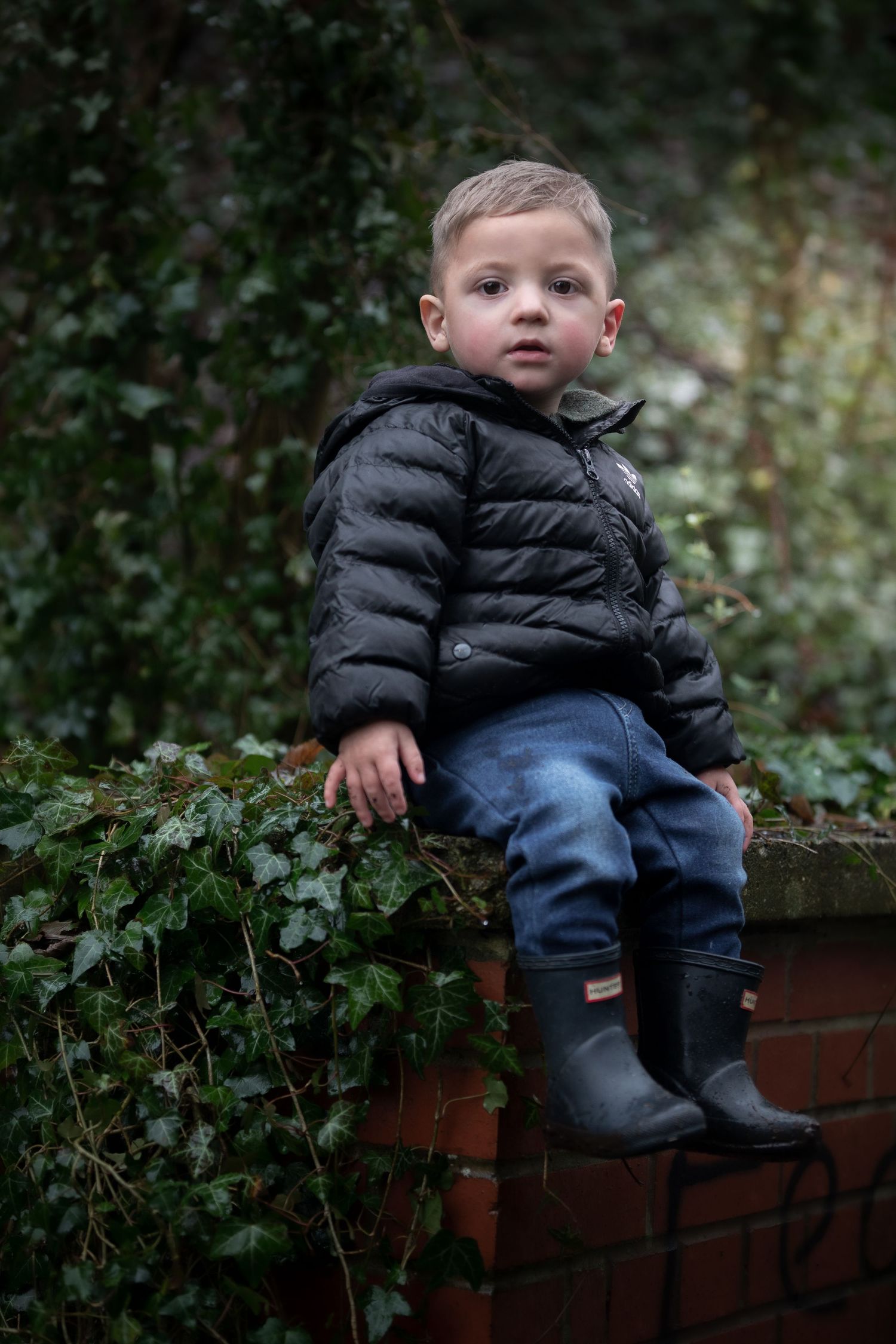 Toddler boy in black puffer jacket and Hunter boots sitting on ivy-covered brick wall outdoors.

Sutton Village