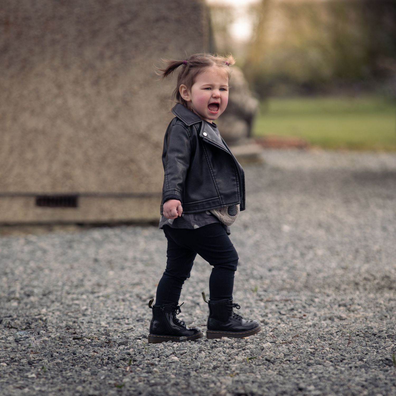 Laughing toddler girl in leather jacket and boots walks on gravel path outdoors.