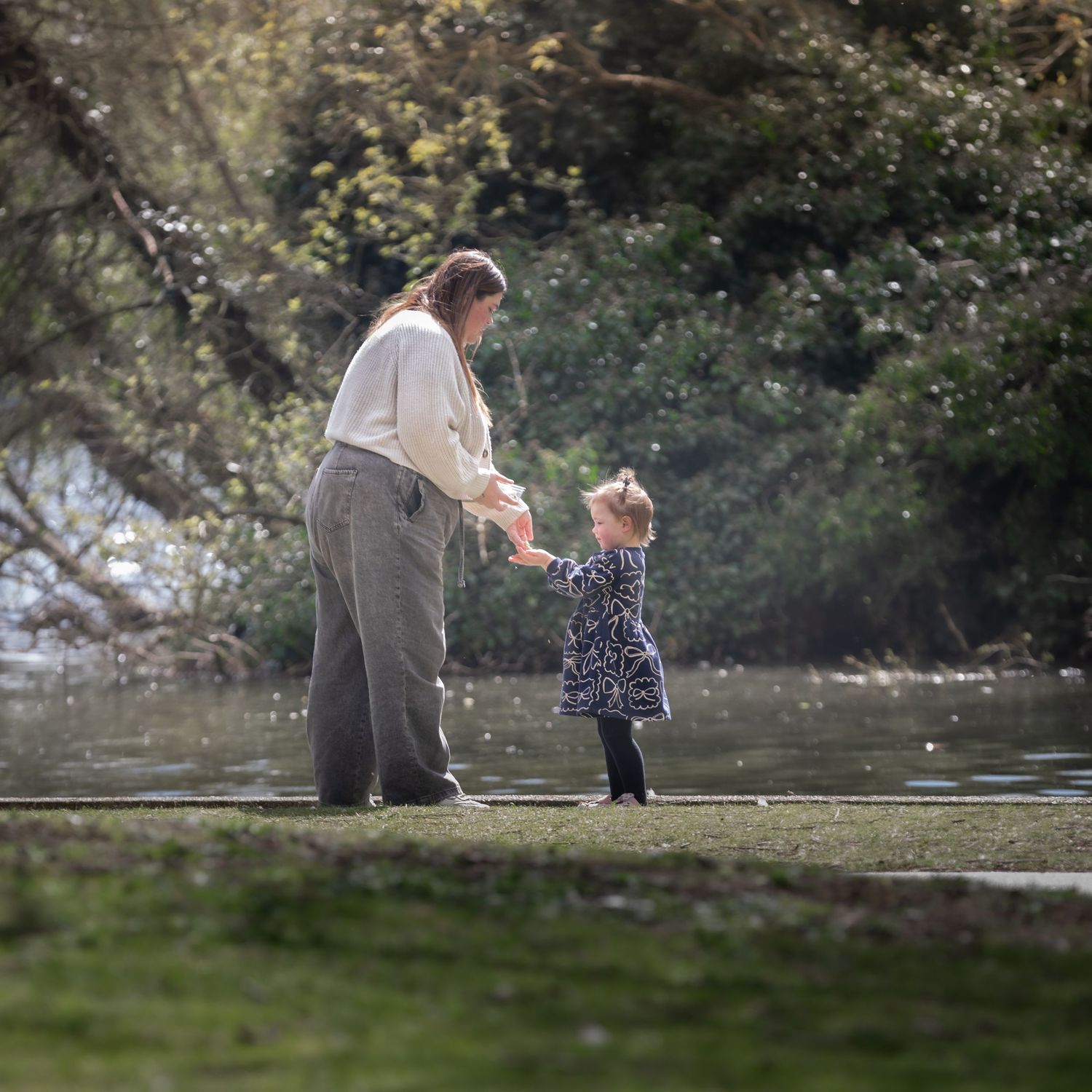Mother and toddler daughter holding hands by a peaceful riverside in sunlight.