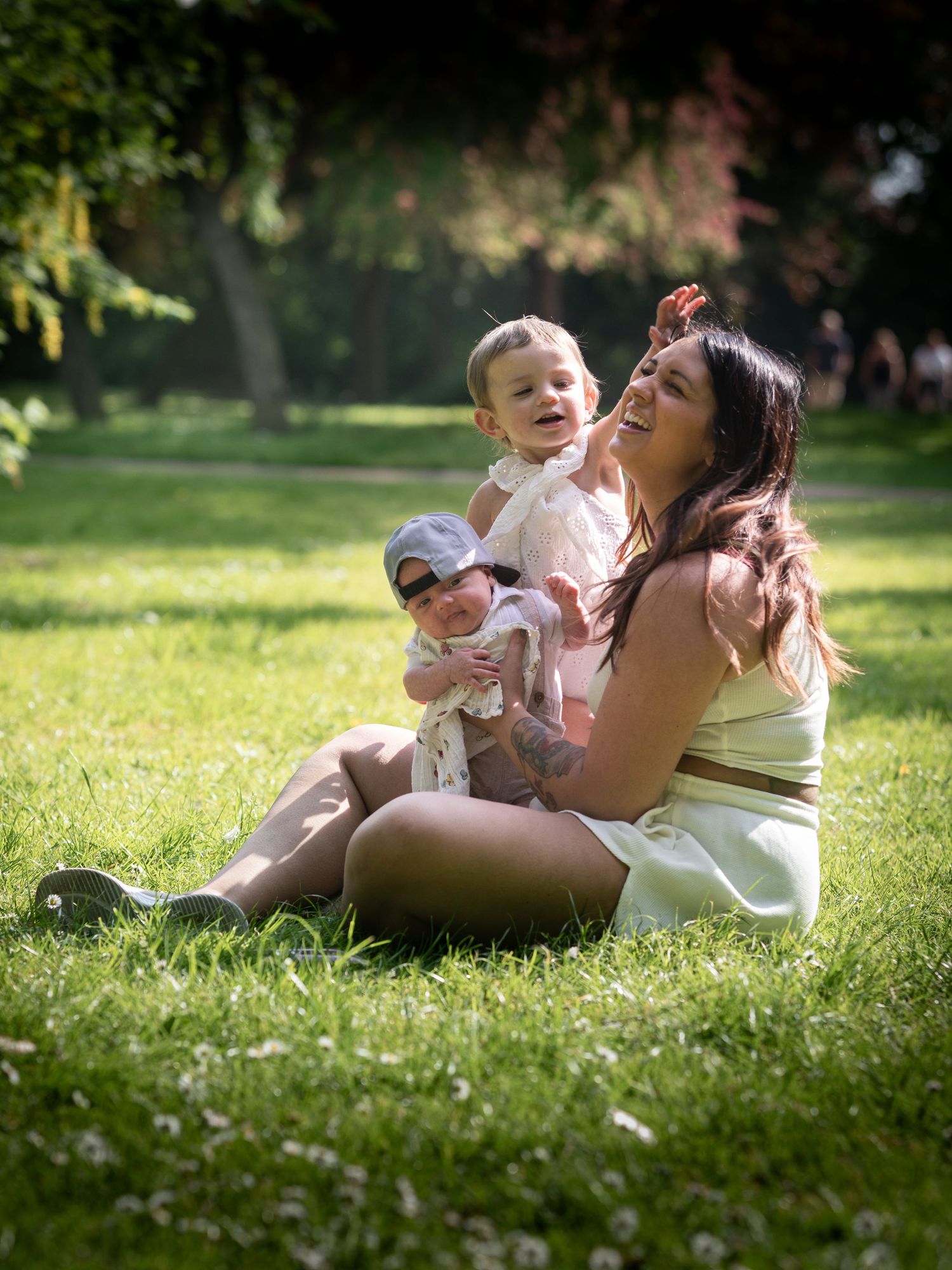 A young mother sits on park grass laughing with her toddler and baby on a sunny summer day.