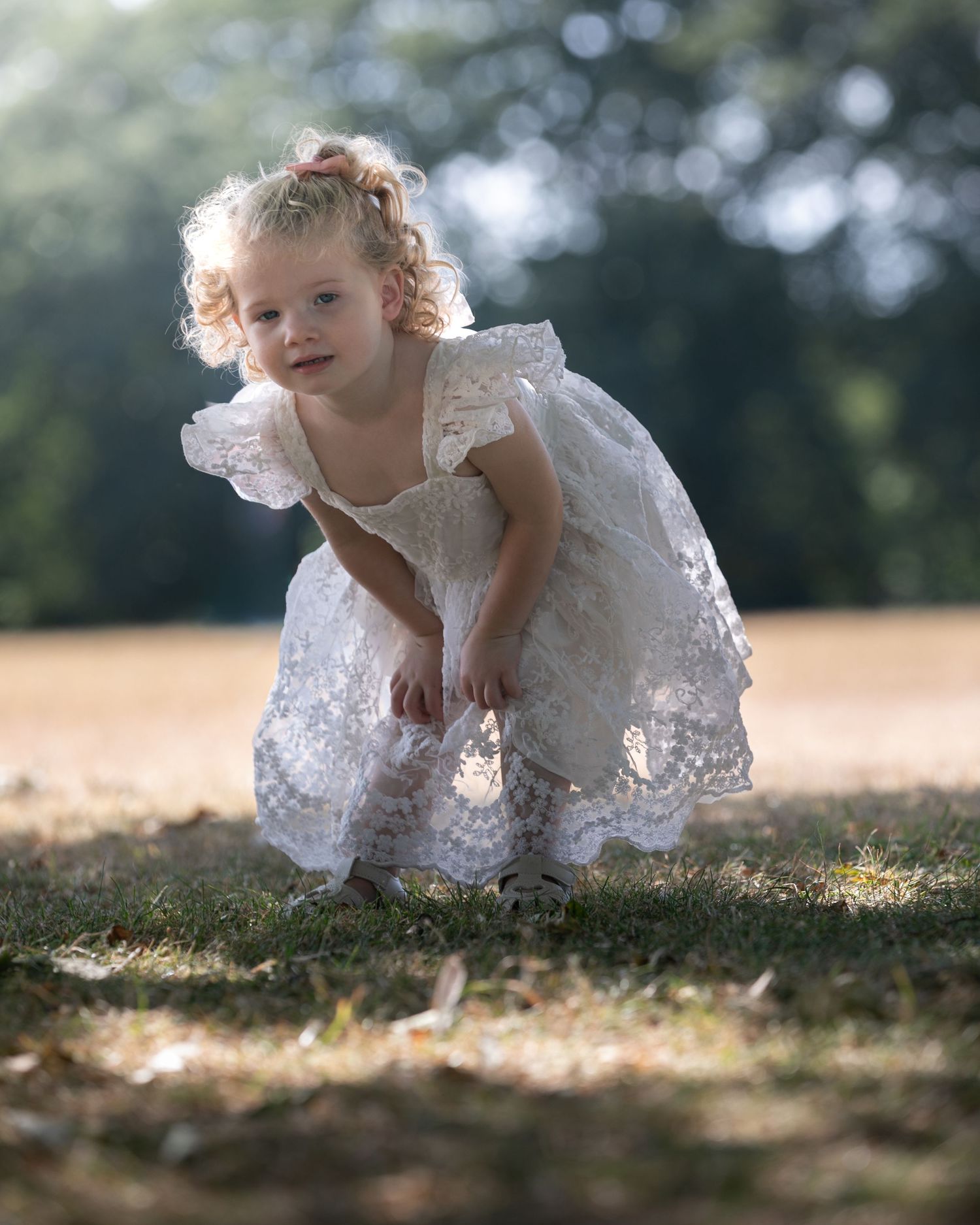 A curly-haired toddler girl in a white lace dress crouches on grass in a sunlit park.