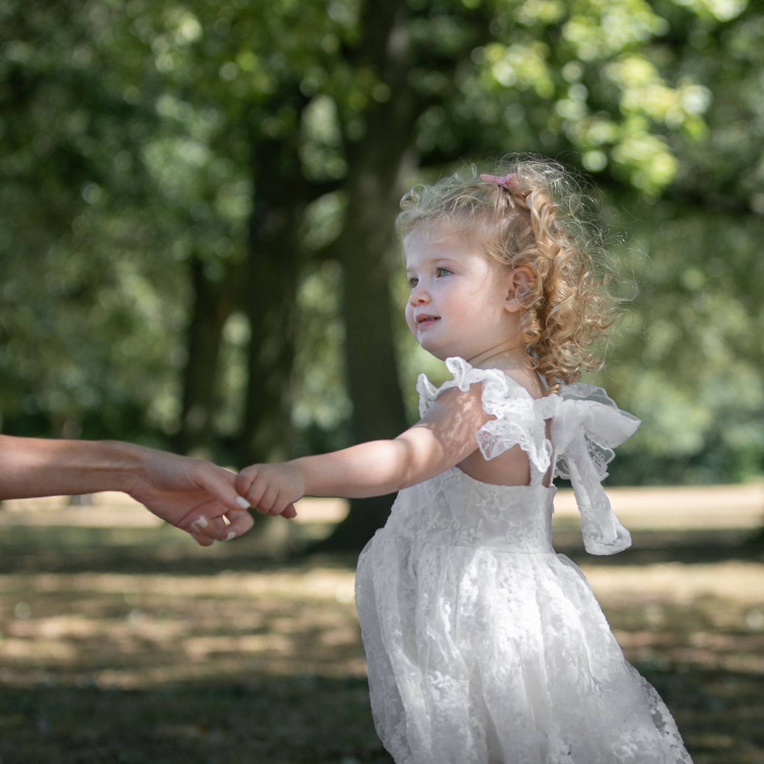 A curly-haired toddler girl in a white lace dress holds an adult's hand in a sunlit green park.