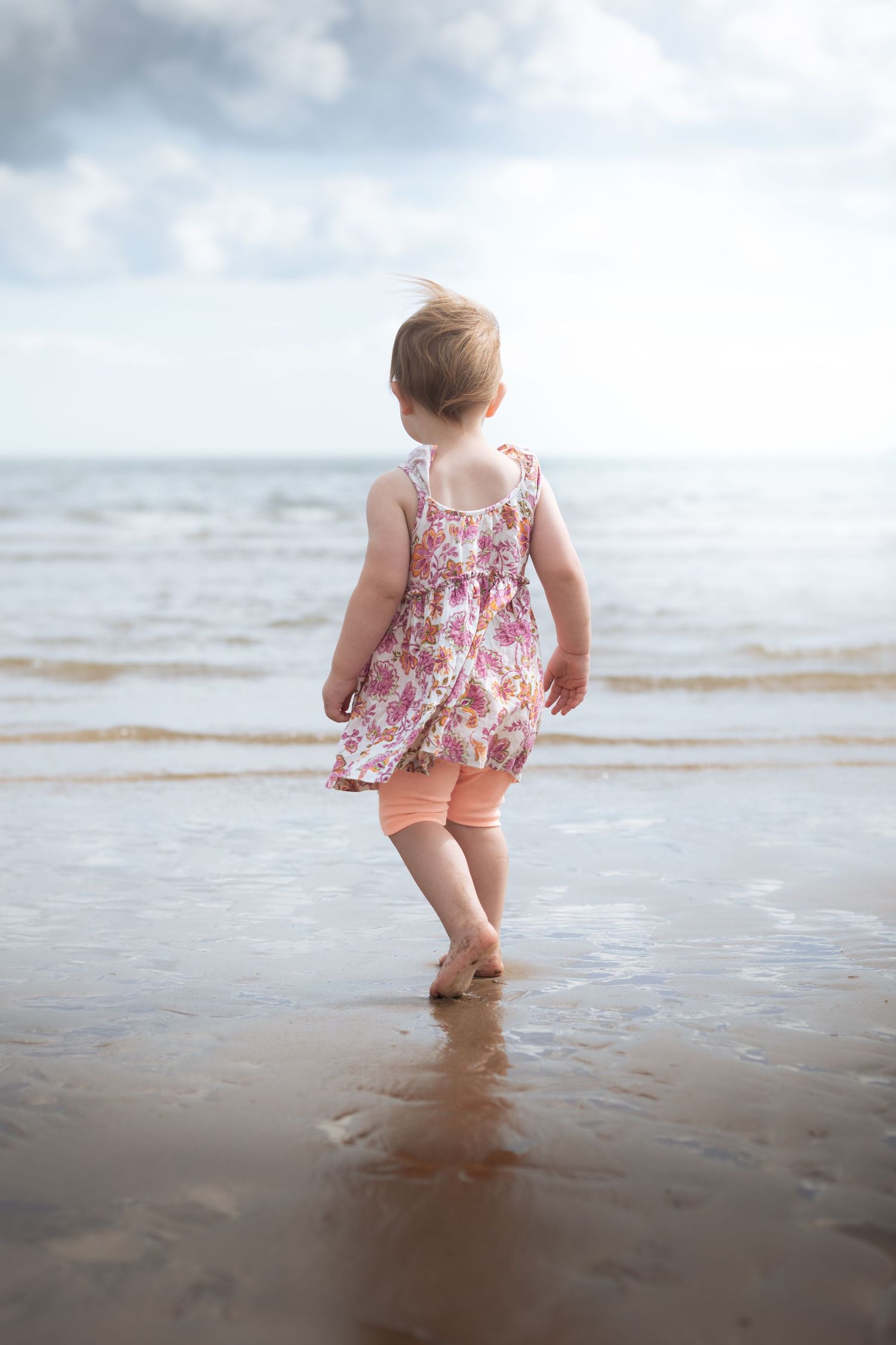 Baby girl in a floral dress walking toward the ocean on a sandy beach.

Scarborough