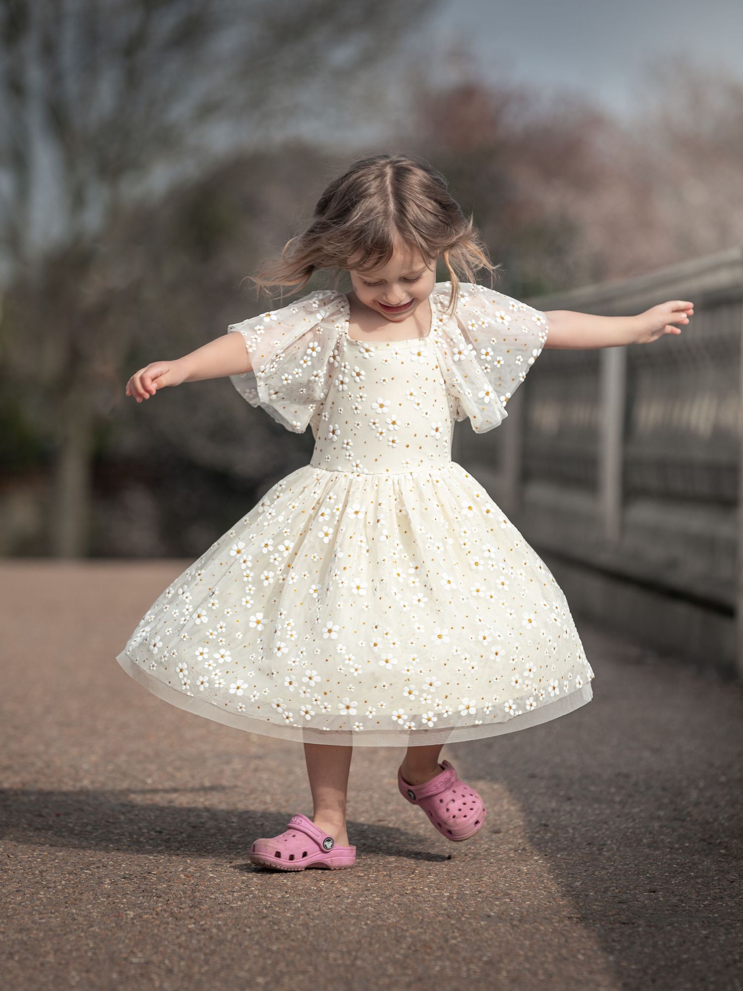 Little girl in cream floral dress twirls joyfully on a path wearing pink Crocs.
East Park Hull
