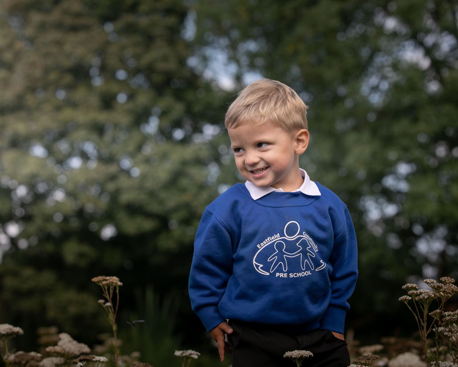 Young boy in royal blue school sweatshirt smiling outdoors with green trees in background.