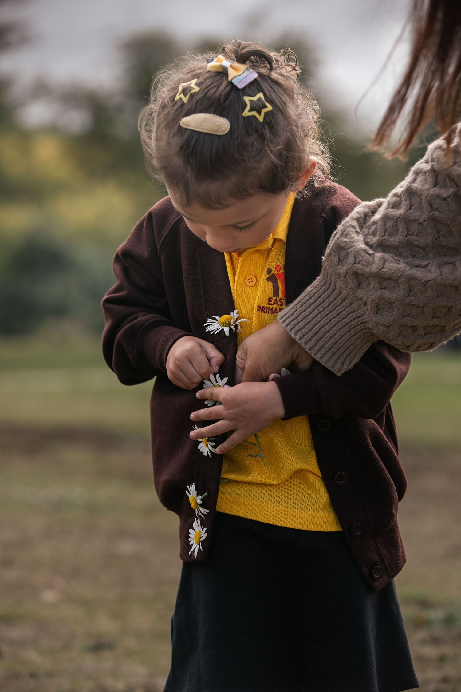Young girl in school uniform with daisy flowers, wearing yellow polo and dark cardigan with star hair clips outdoors.