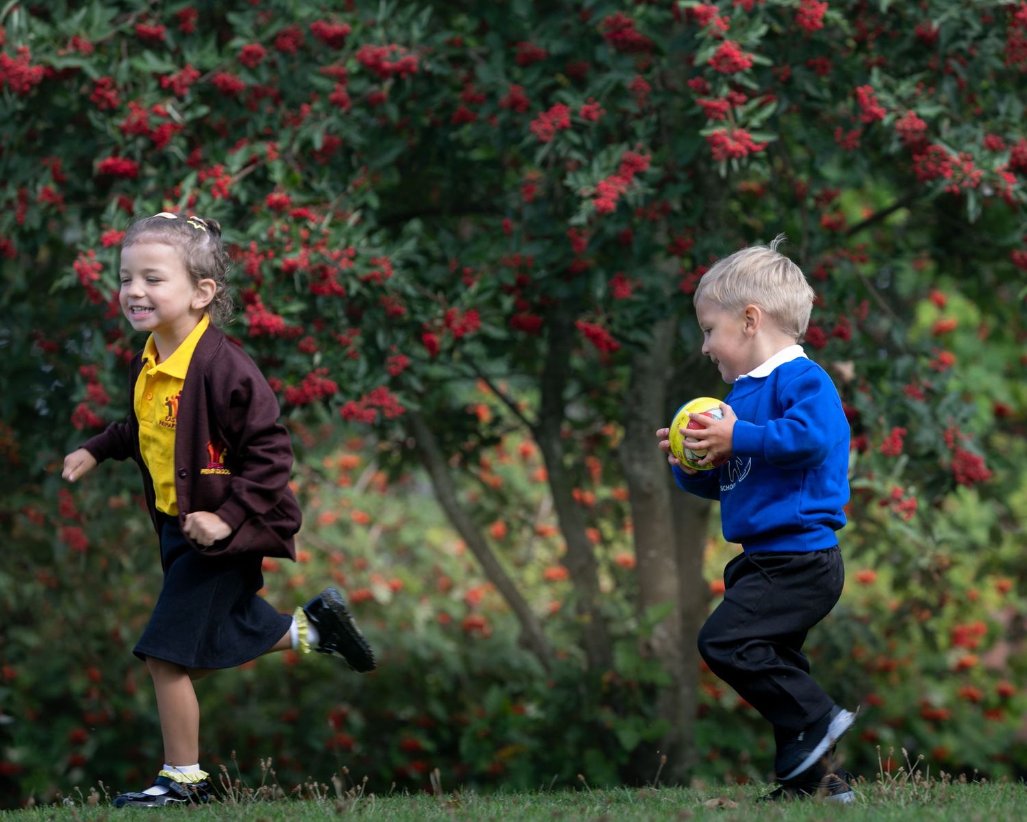 Two young children in school uniforms running outdoors near a red berry tree on green grass.