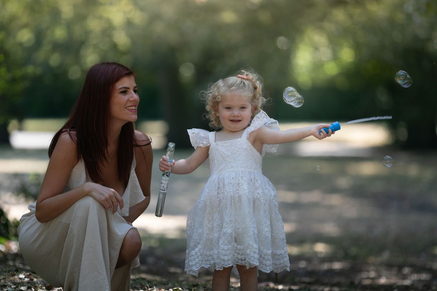 A mother and toddler girl in white dress playing with bubbles outdoors in a sunny green park.