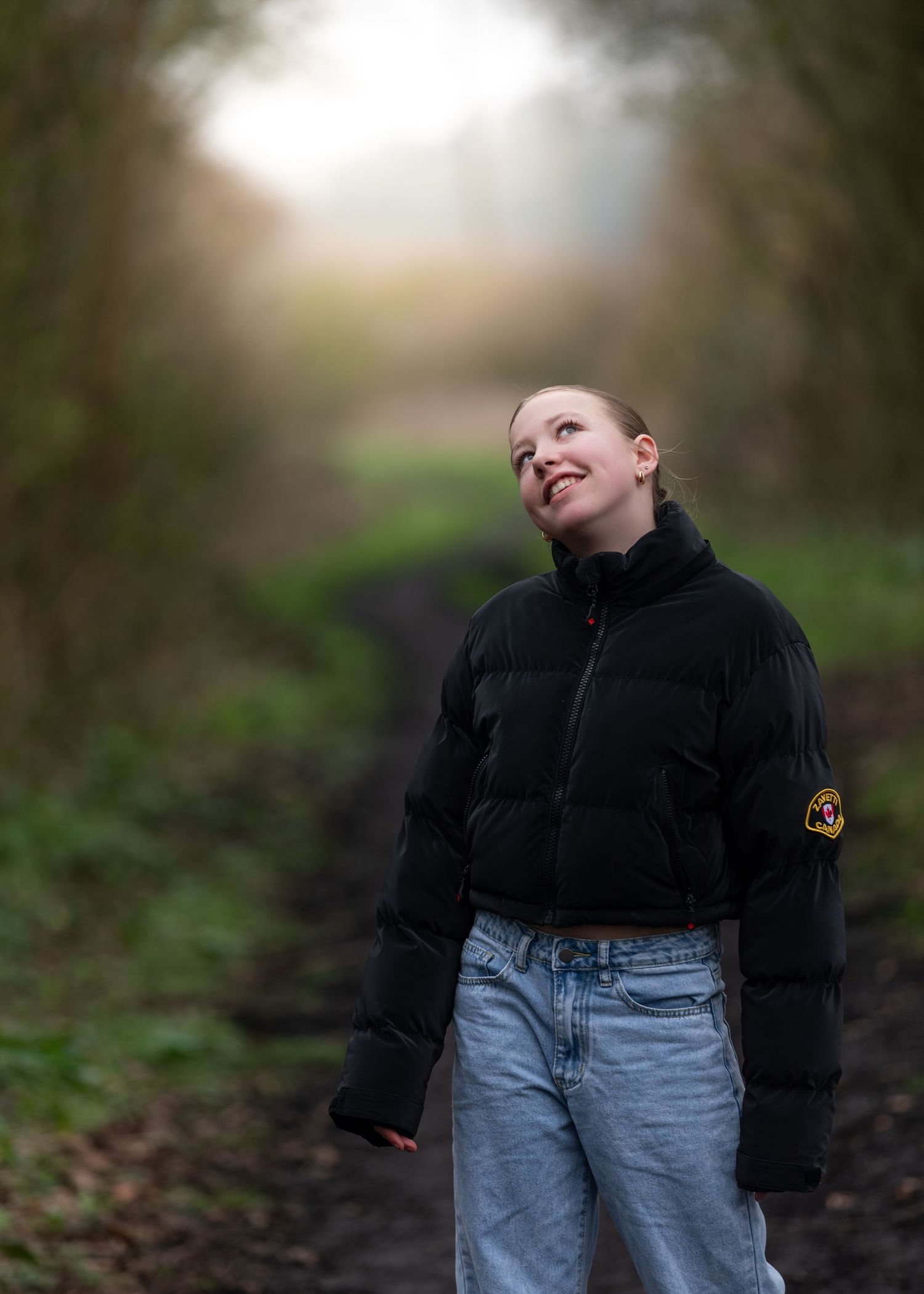 Young woman in black puffer jacket and jeans smiling on misty woodland path in natural light.

Keyingham