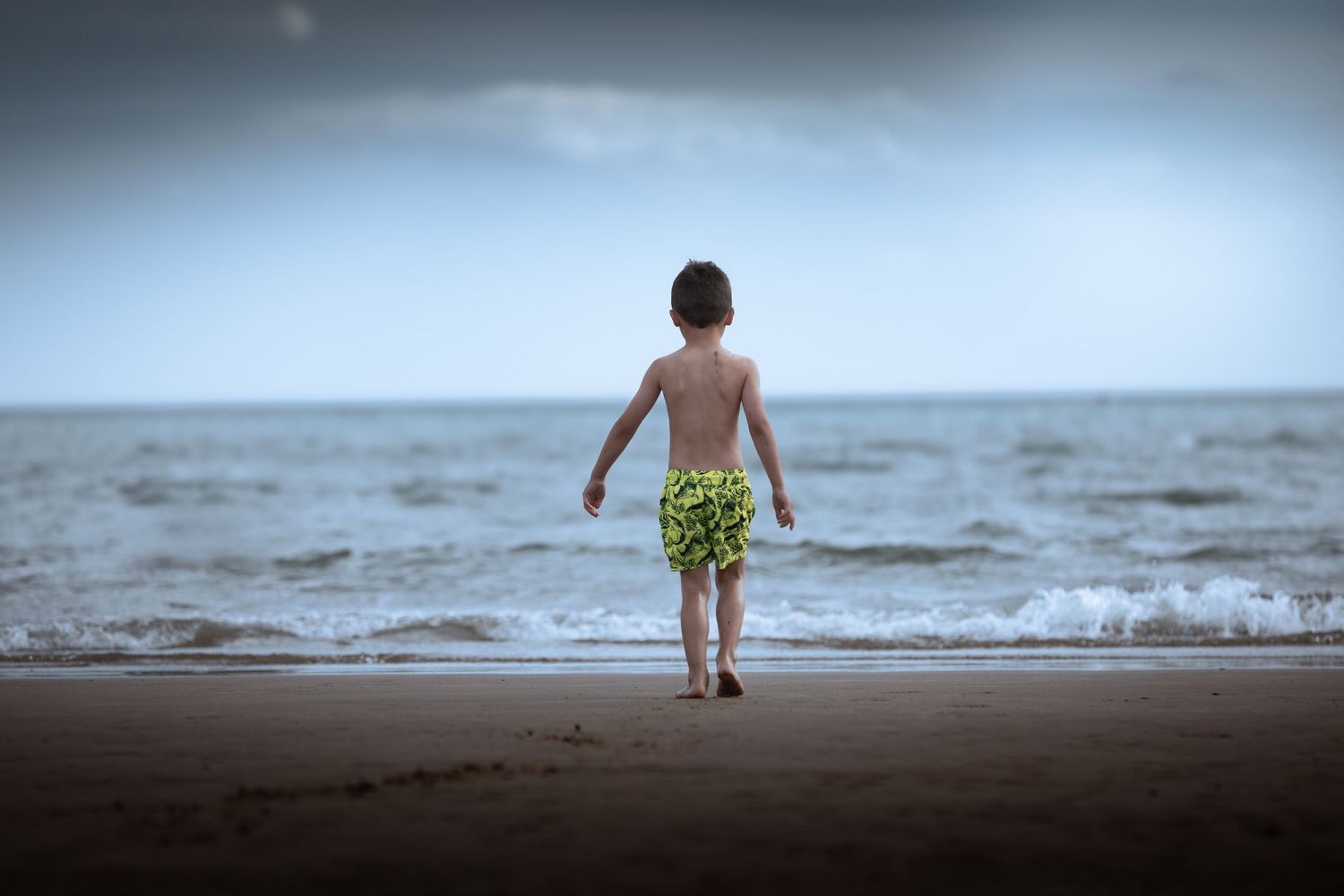 Young boy in green swim shorts walking toward ocean waves on a cloudy beach.

Scarborough