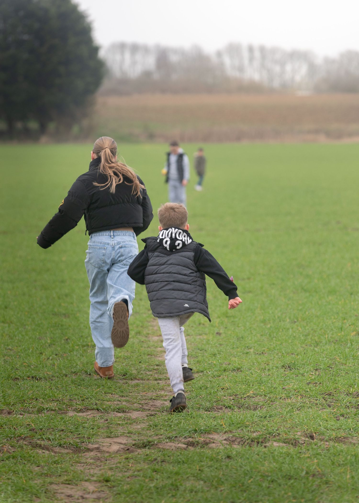 A woman and young child run together across a wide green field on an overcast day in the countryside.