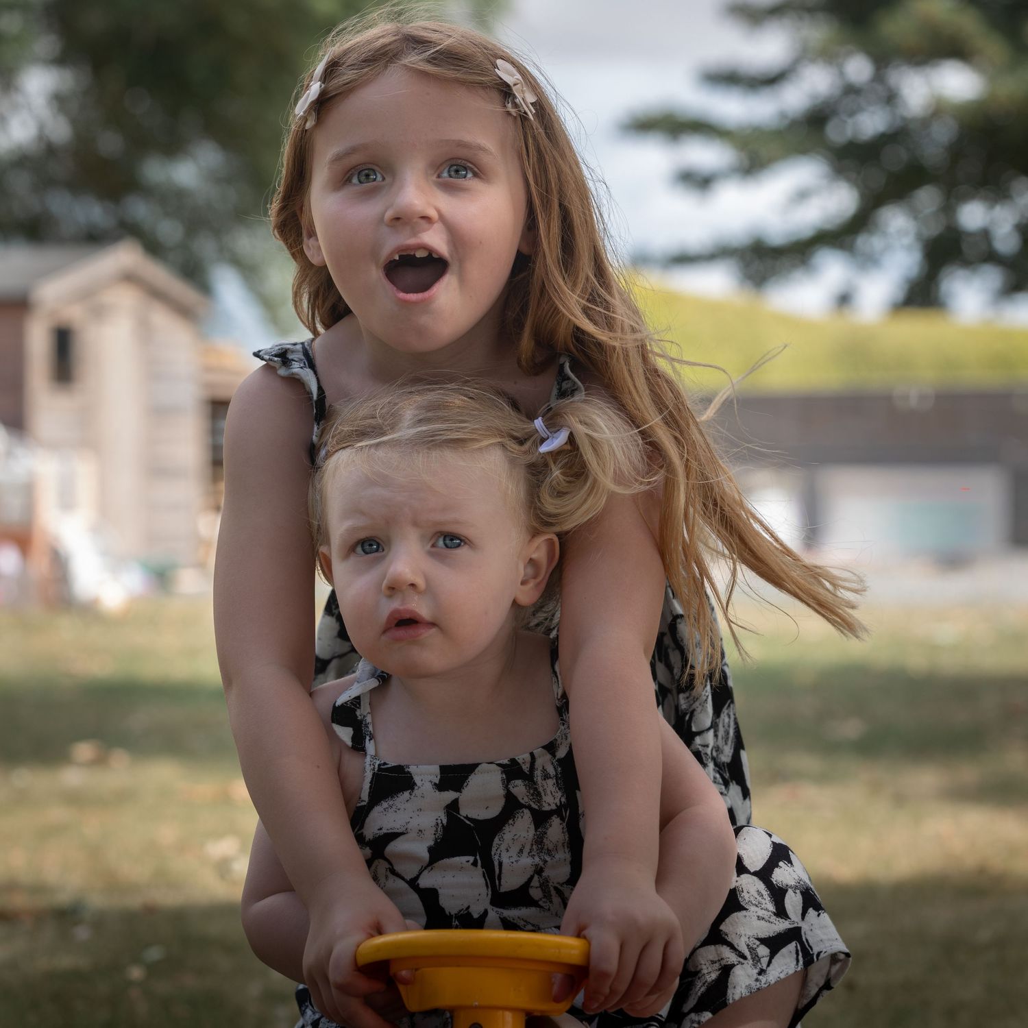 Two young girls in matching floral dresses play outdoors, the older girl hugging her toddler sister with a surprised expression.