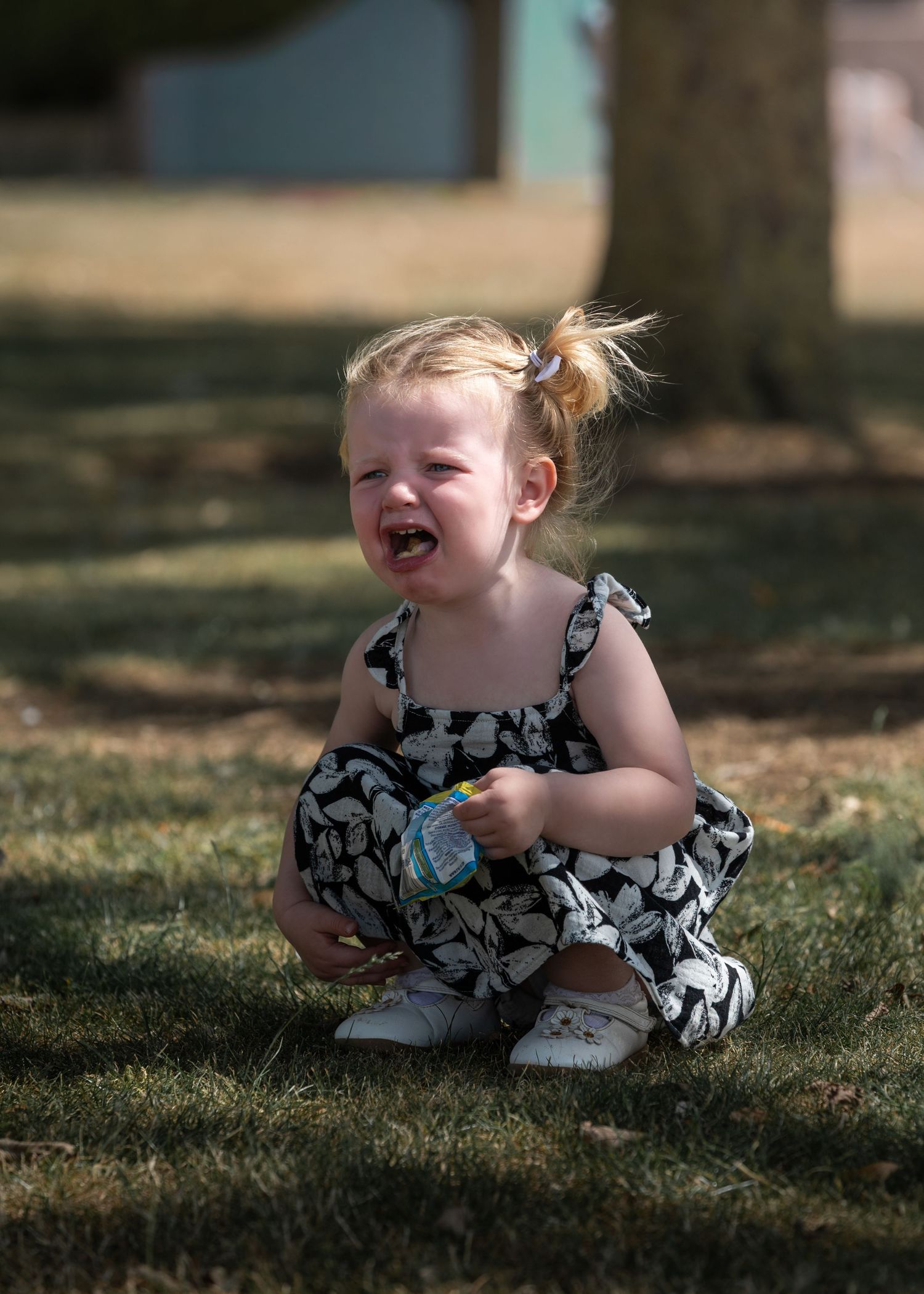 Smiling toddler girl in black and white floral romper sitting cross-legged on grass outdoors on a sunny day.