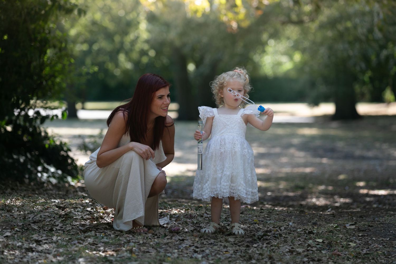 A woman crouches beside a curly-haired toddler in a white dress holding a flower in a sunny park.