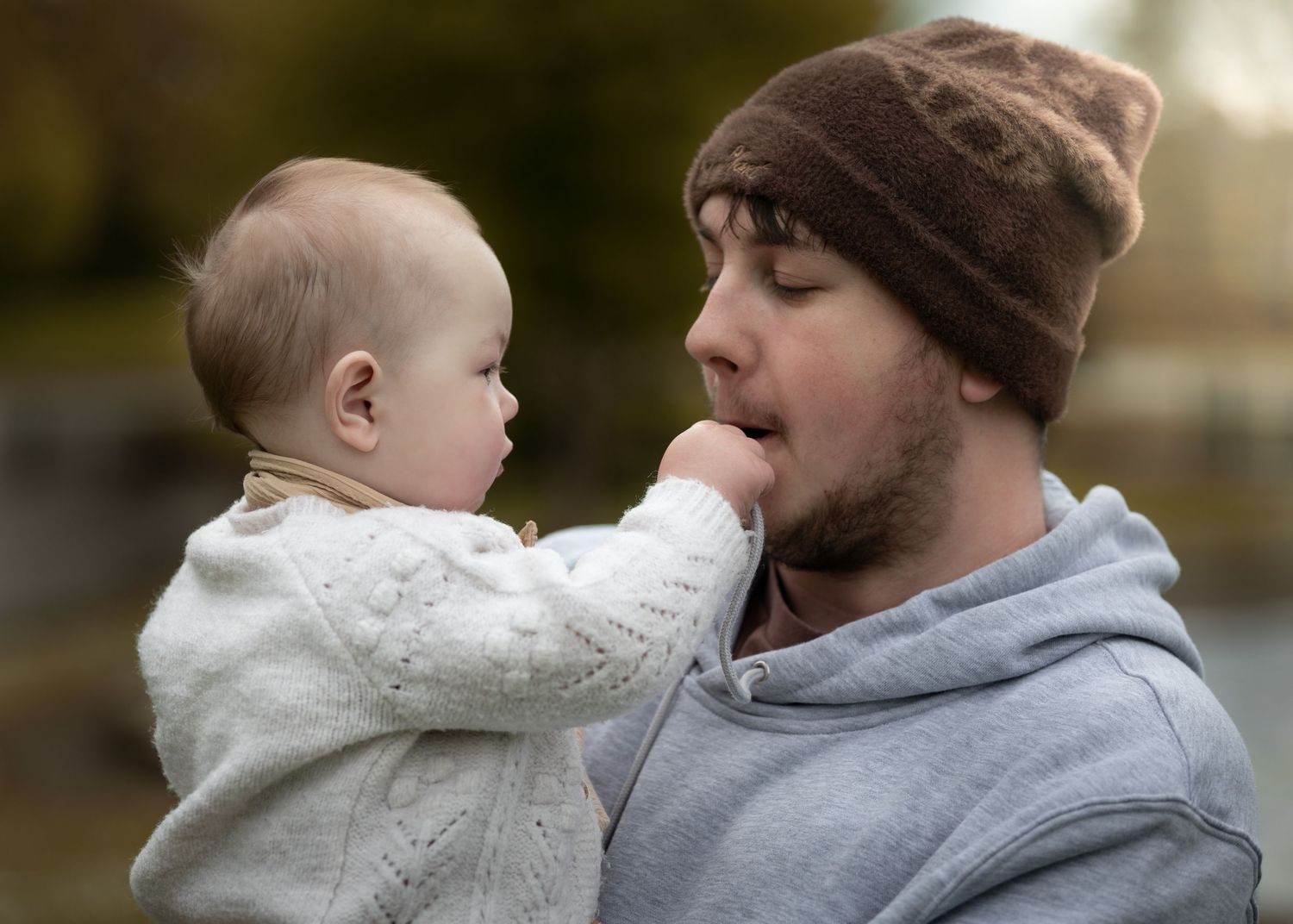 A bearded man in a brown beanie holds a baby who reaches toward his mouth in a tender outdoor moment.