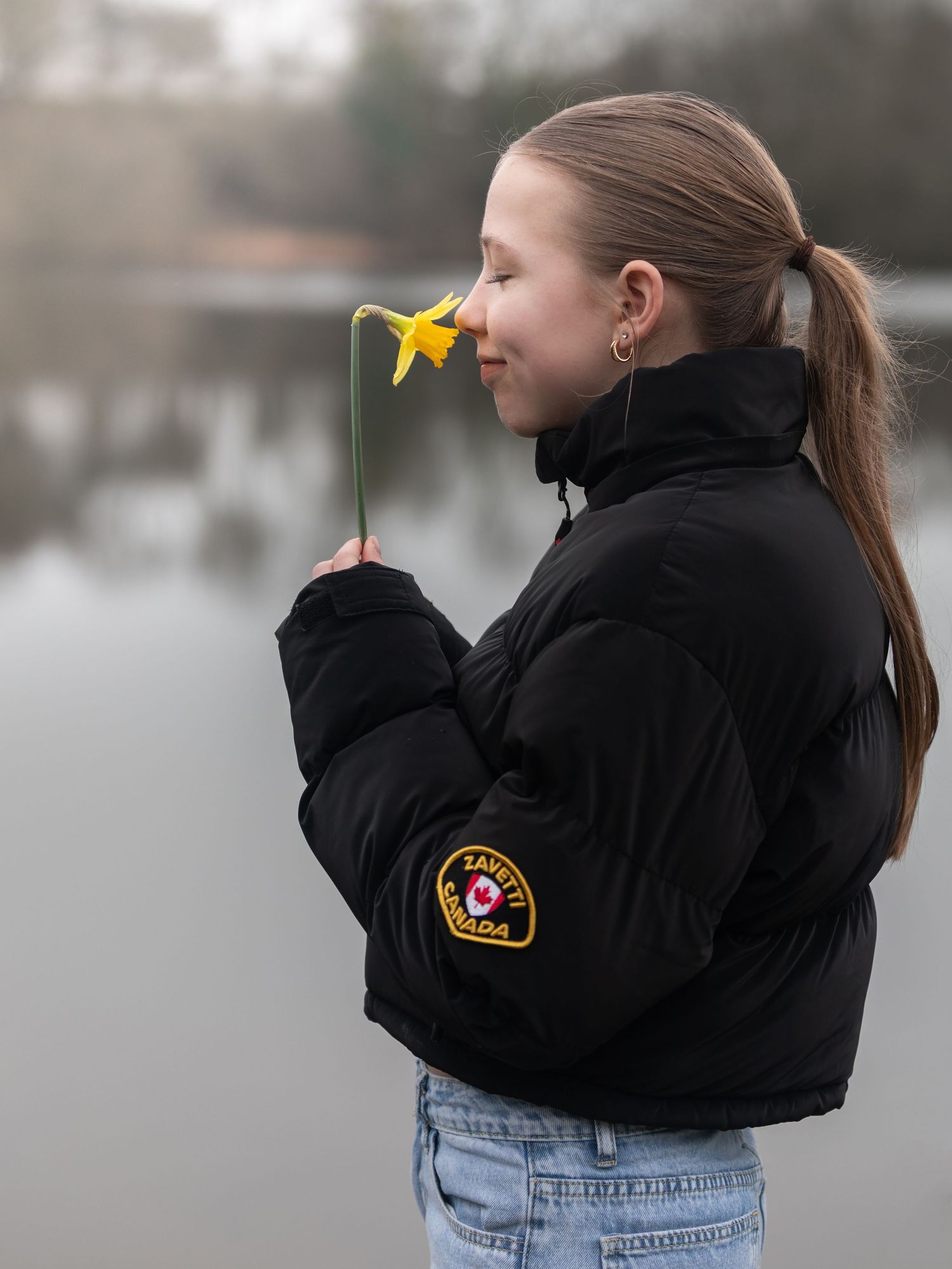 A teenage girl in a black puffer jacket smells a yellow daffodil beside a calm misty lake.

Keyingham