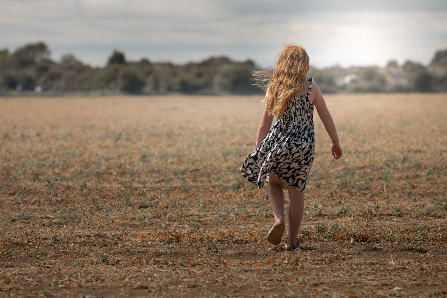 Young girl in floral dress walking through a golden harvested field with trees on the horizon.