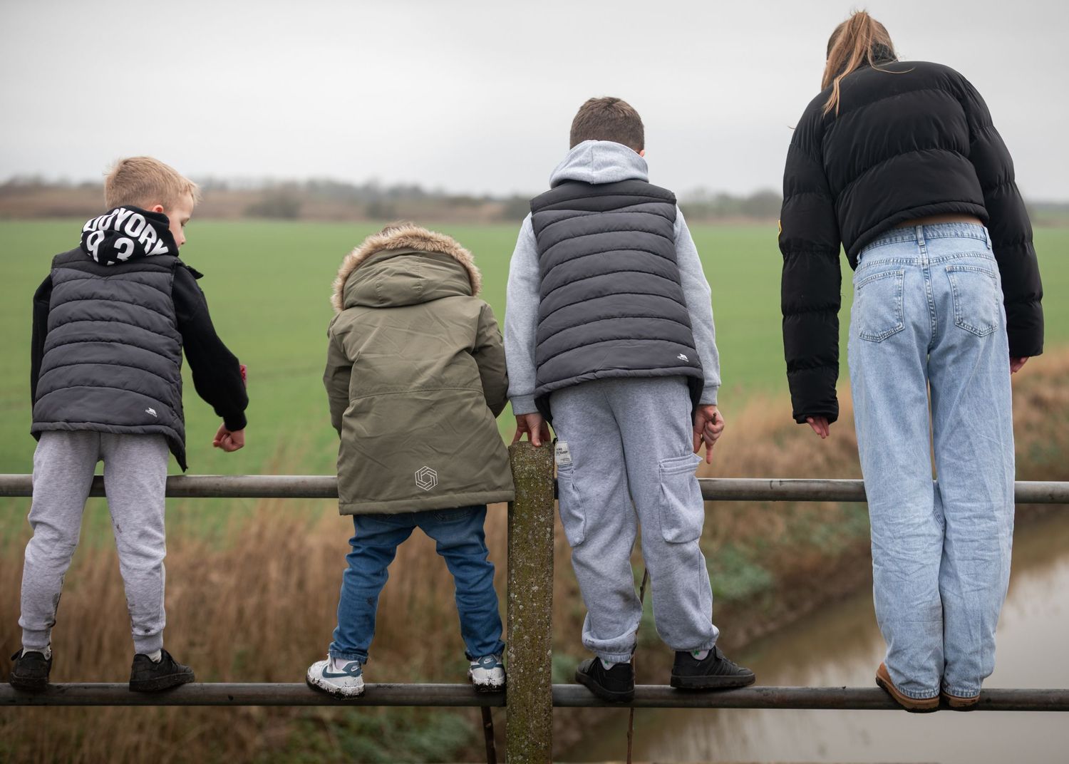 Four children stand on a fence railing viewed from behind, overlooking a green field on an overcast day.

Keyingham