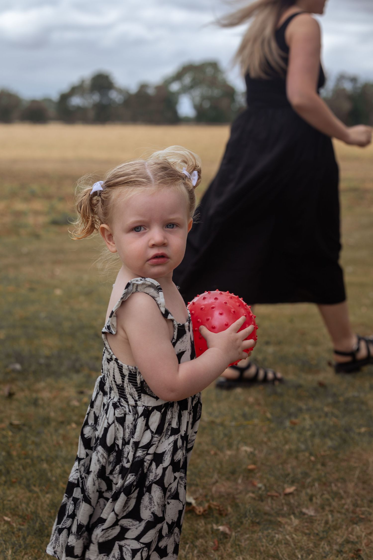 Toddler girl in patterned dress holding red ball in open field, with woman in black dress in background.